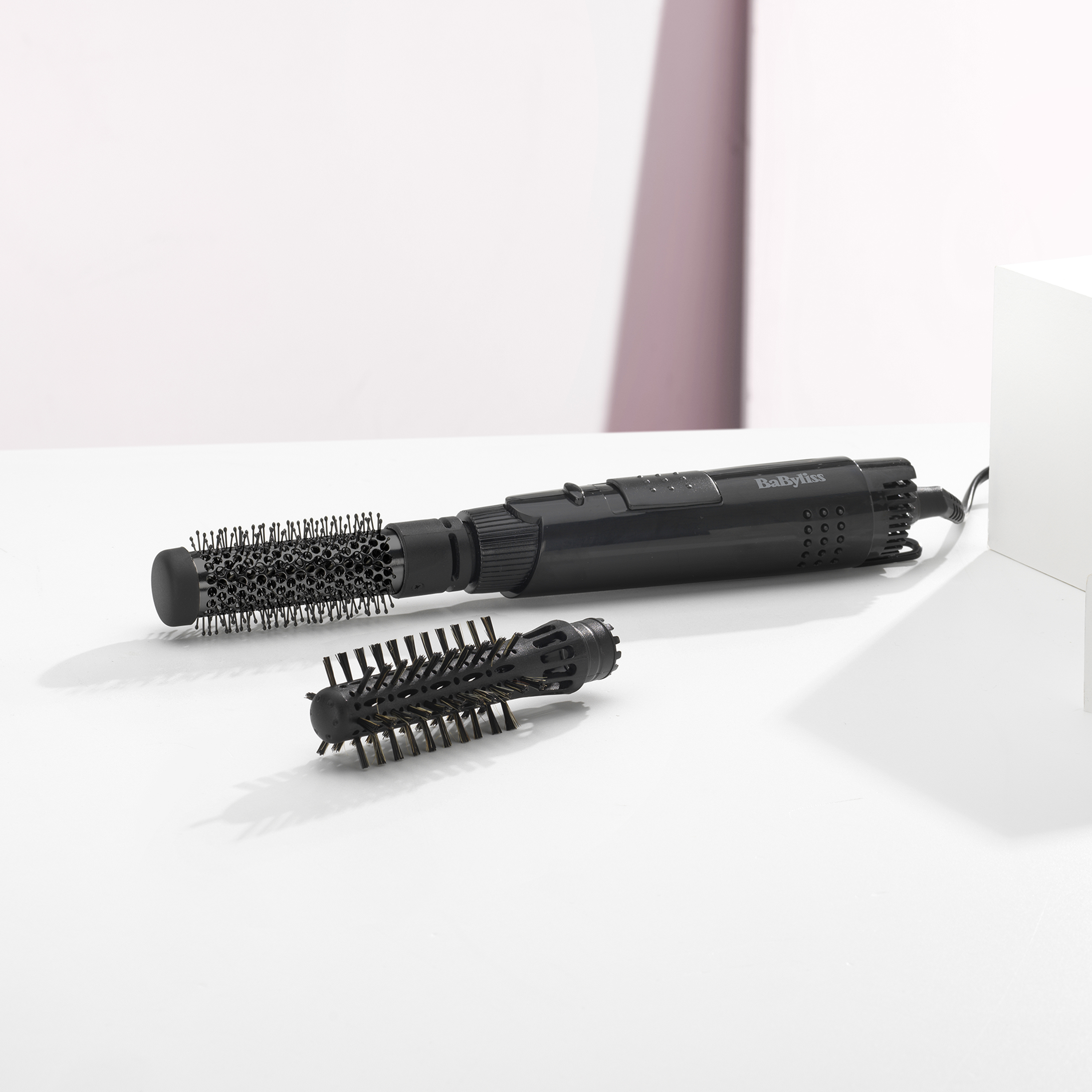 Ein schwarzer Babyliss-Haarstyler mit zwei Aufsätzen auf weißer Oberfläche mit weißem und rosa Hintergrund.