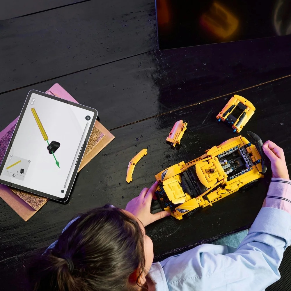 Dziecko składa żółty samochód Lego z tabletem pokazującym instrukcje. Części samochodu leżą na czarnym stole.