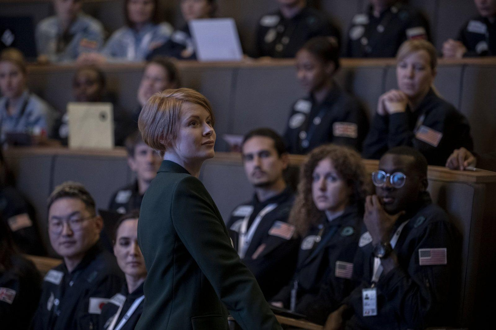 Eine Frau in einem grünen Blazer blickt in einem Hörsaal nach oben. Im Hintergrund sitzen Menschen in dunklen Uniformen, einige mit US-Flaggen.