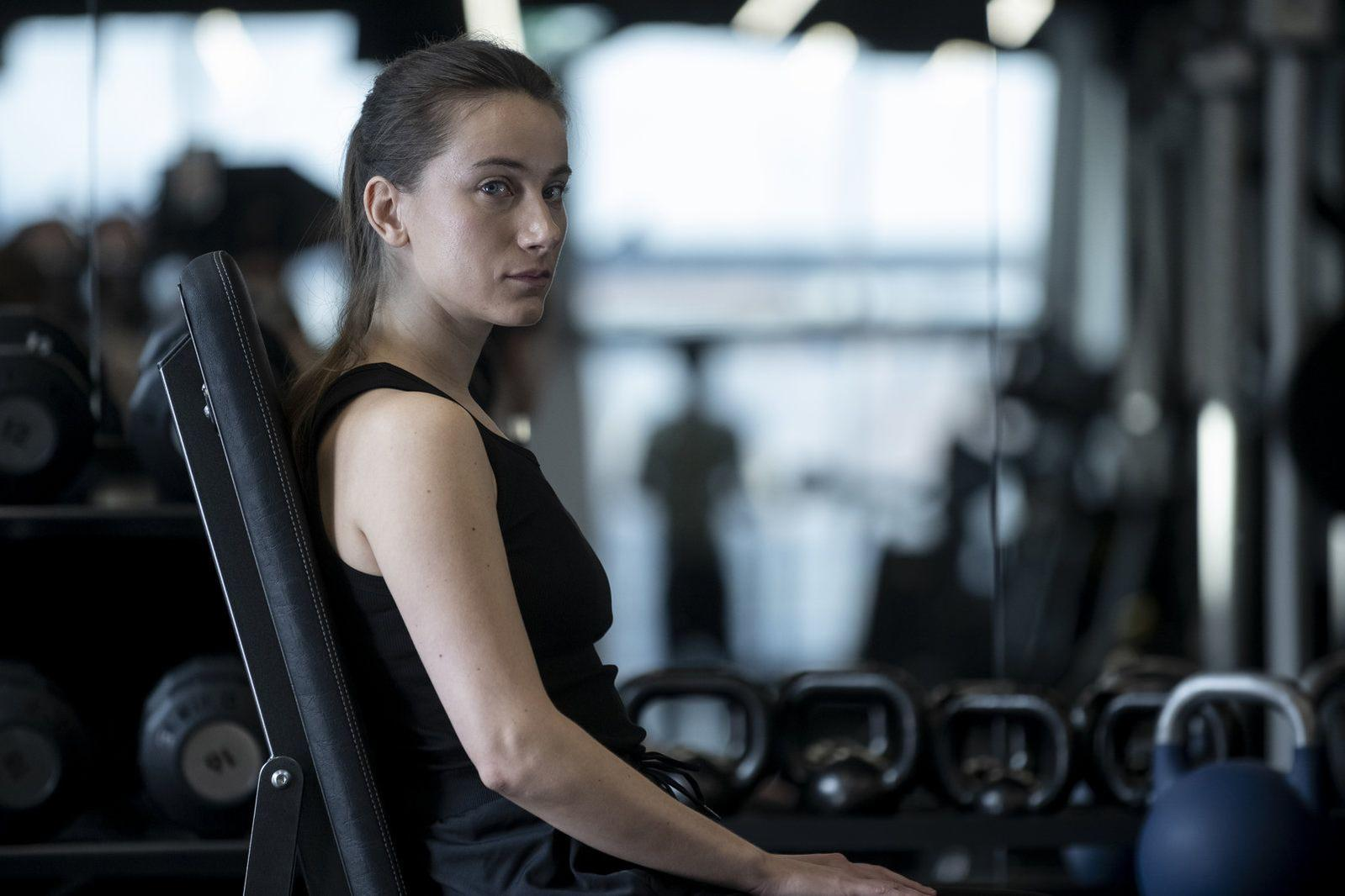 Eine Frau in einem schwarzen Tanktop sitzt auf einer Hantelbank in einem Fitnessstudio und schaut in die Kamera. Gewichte sind im Hintergrund.