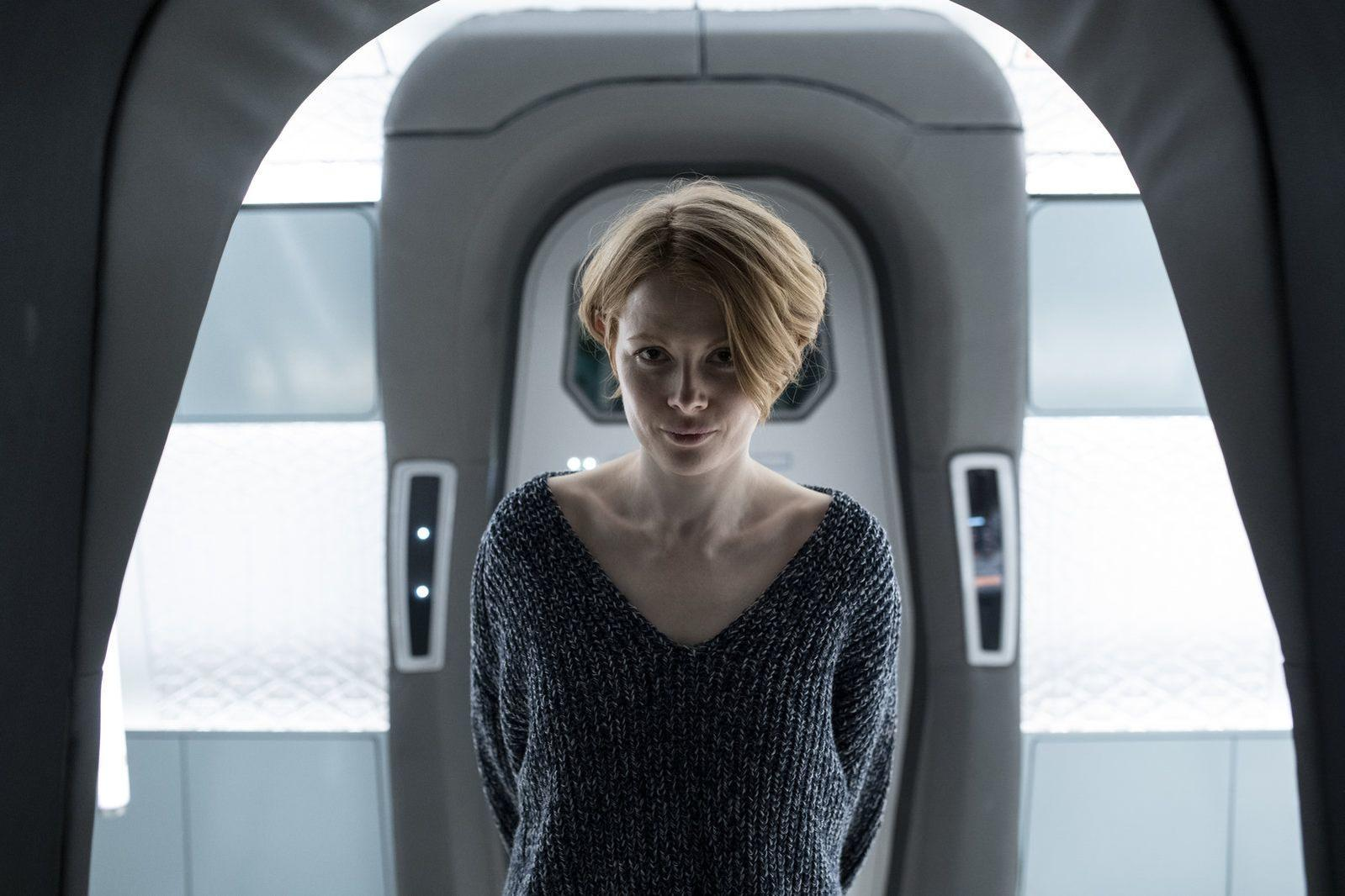 Eine Frau mit kurzen blonden Haaren trägt einen grauen Pullover. Sie steht in einem futuristischen, grauen Raum und schaut in die Kamera.