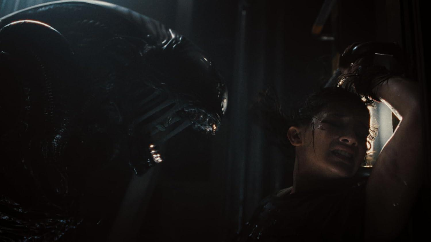 Ein außerirdisches Wesen ragt über einer verängstigten Person in dunkler Umgebung. Das Alien hat einen langen Kopf und einen bedrohlichen Blick.