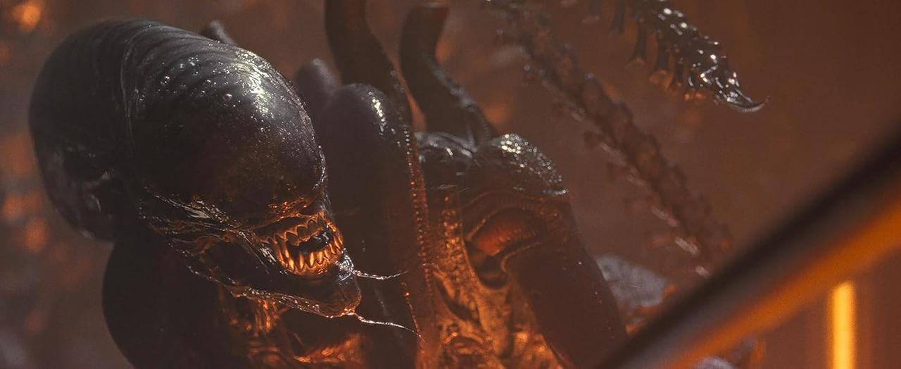 Ein Alien-Kreatur mit einem dunklen, strukturierten Kopf und scharfen Zähnen. Die Szene ist schwach mit Orangetönen beleuchtet.