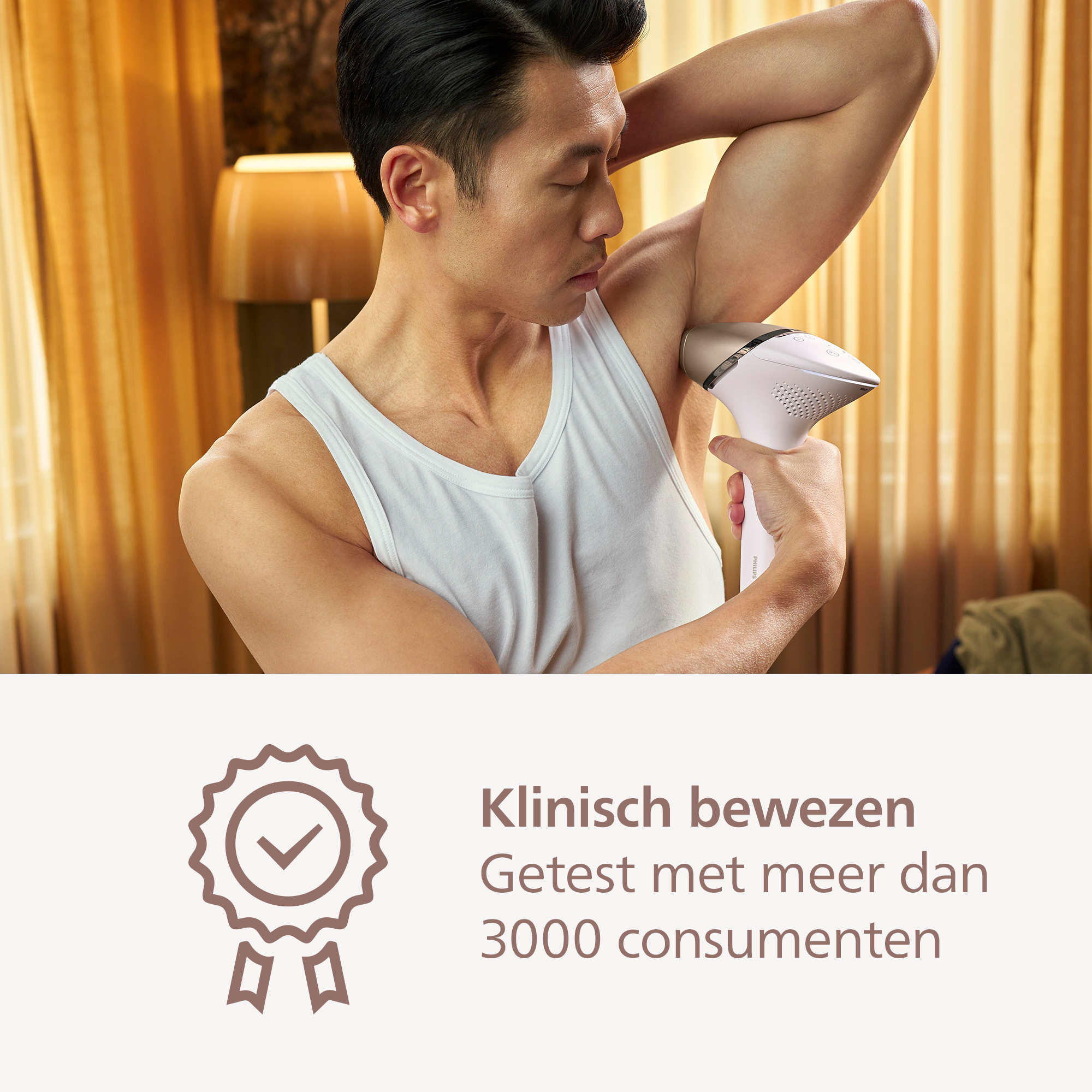 Man using a hair removal device on his armpit. He wears a white tank top. Background has curtains and a lamp.