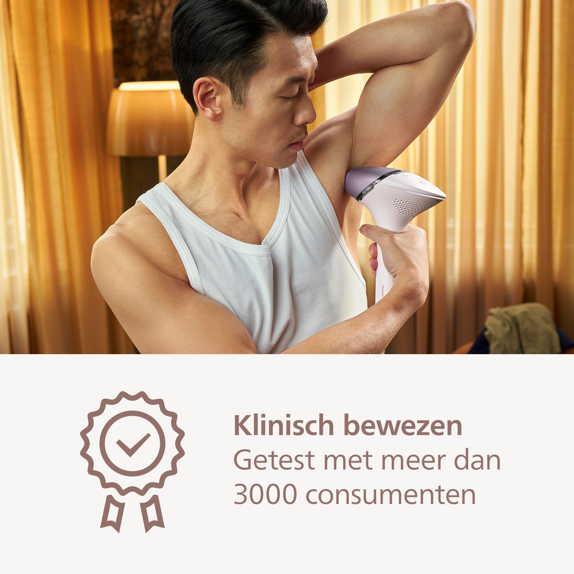 A man in a white tank top uses a hair removal device on his armpit. Background includes curtains and a lamp.