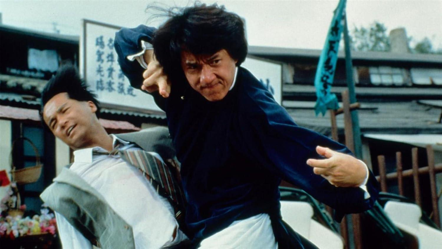 Jackie Chan in einer Kampfszene. Er schlägt einen Mann im Anzug, der zurückweicht. Im Hintergrund sind Gebäude und eine Flagge zu sehen.
