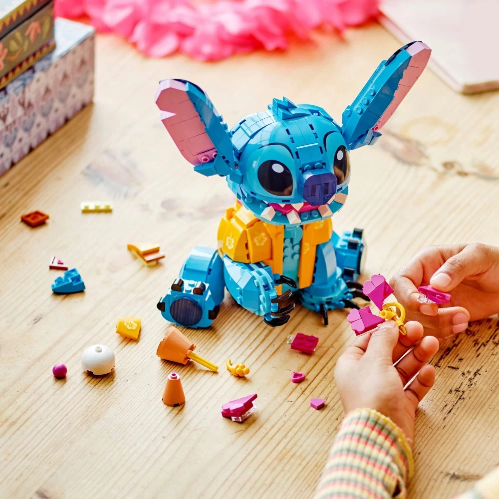 Figurka Lego Stitch jest otoczona elementami, dłonie składają różowe akcesorium. Drewniana powierzchnia, różowa girlanda kwiatowa w tle.