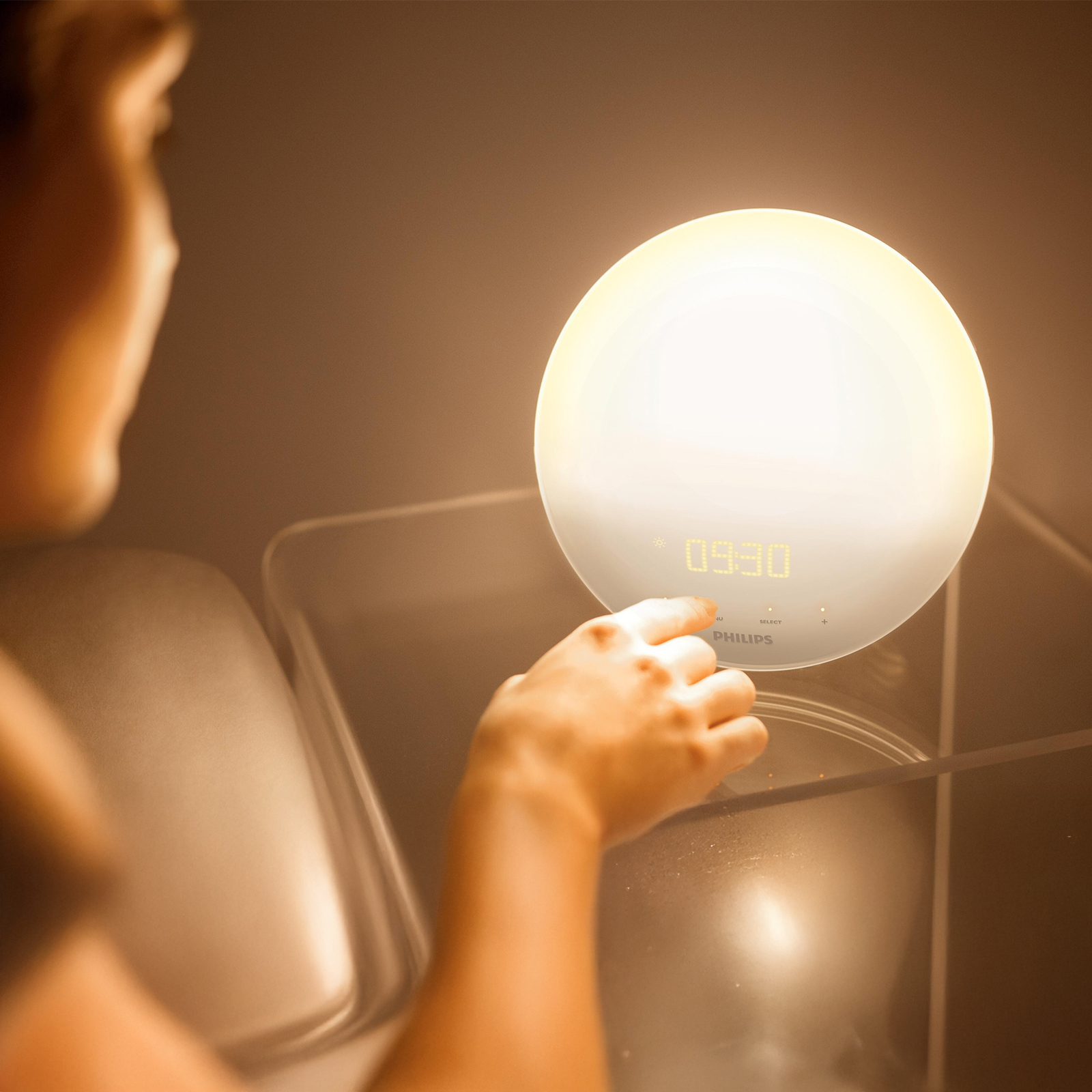 A person's hand touching a white Philips wake-up light. The time 09:30 is displayed. The light is glowing.