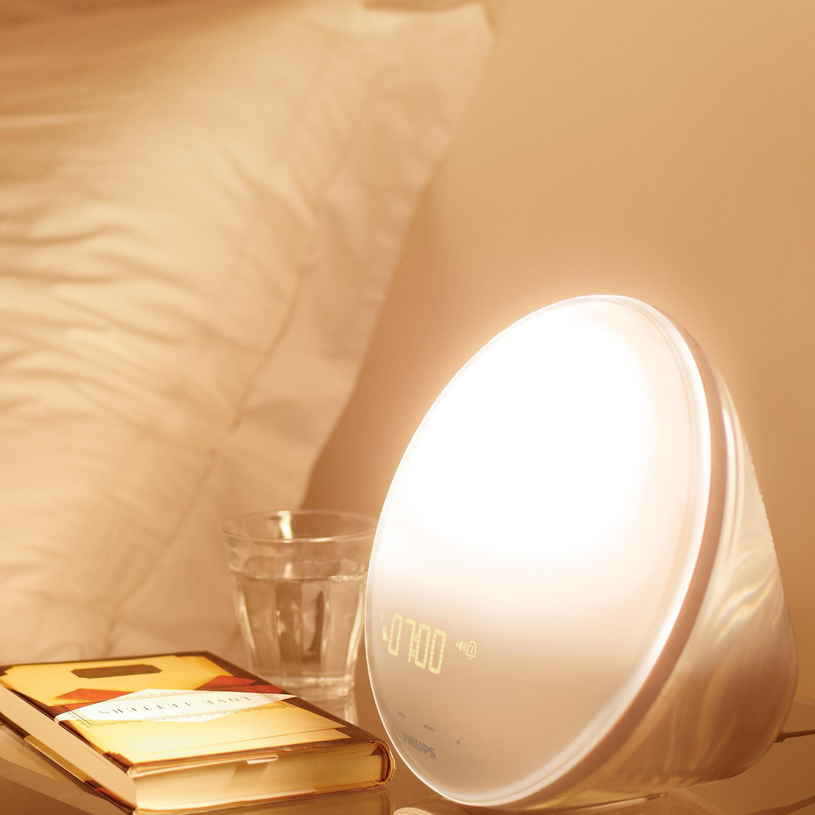 A white Philips wake-up light displays 07:00. Beside it, a glass of water, a book, and a bed.