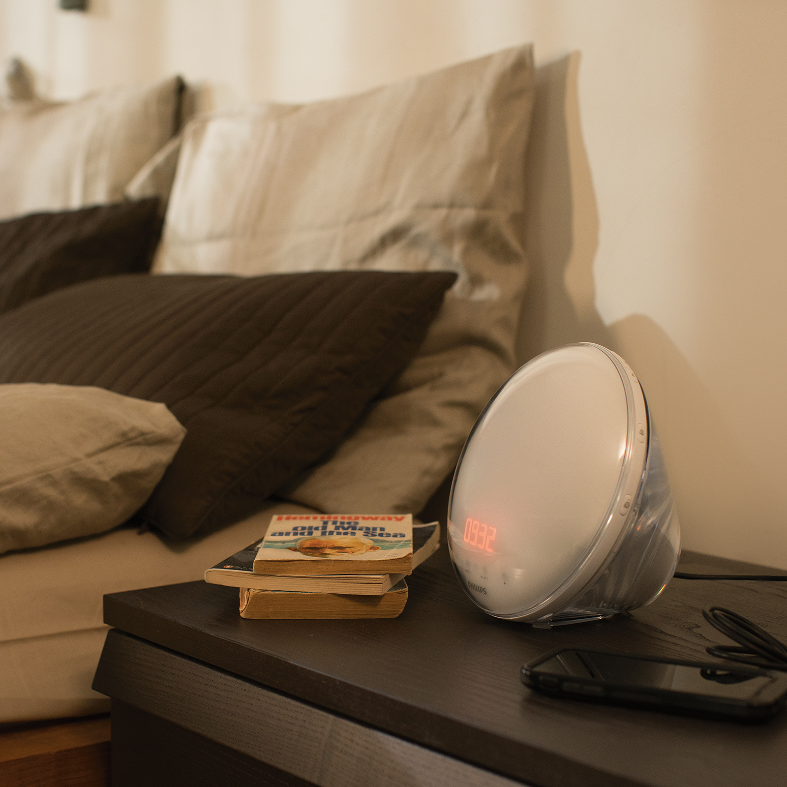 A bedside scene with a Philips alarm clock, books, and a phone. Background features pillows and a bed.