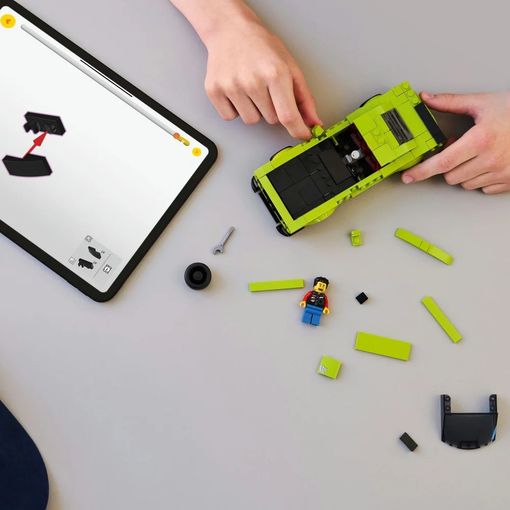 Ręce składają zielony samochód Lego z czarnymi detalami. Rozrzucone części i minifigurka na szarej powierzchni. Tablet pokazuje instrukcje.