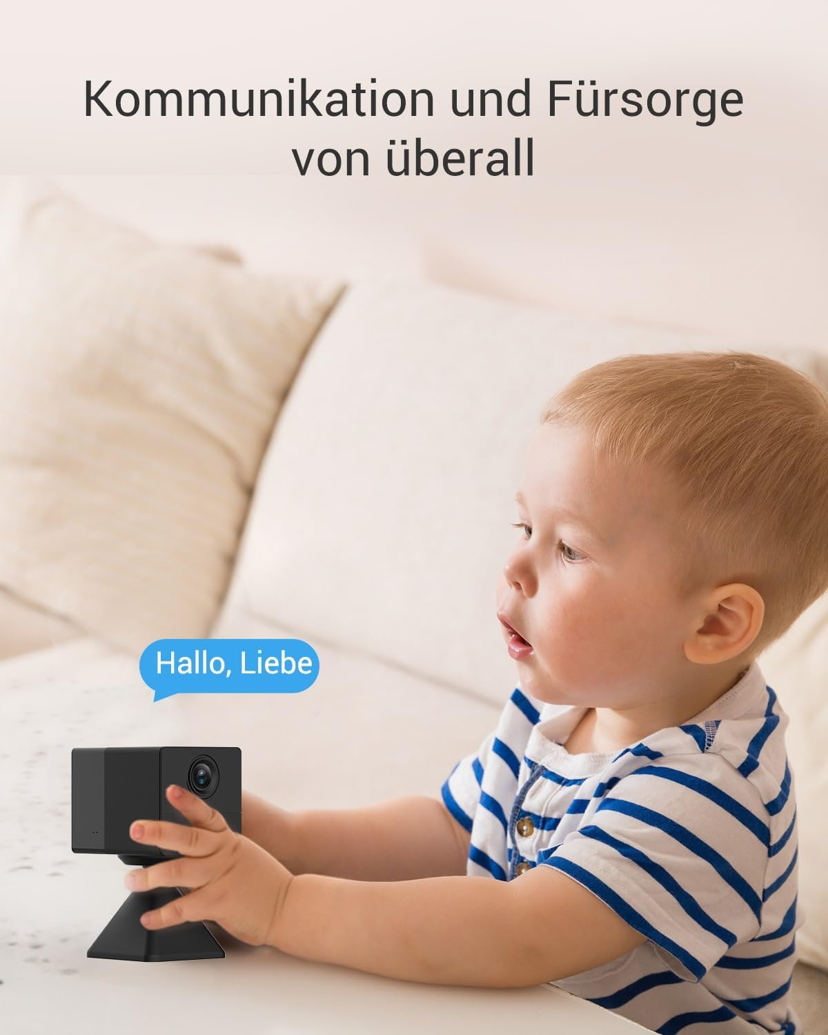 Un bambino guarda una fotocamera nera su un tavolo bianco. Una nuvoletta dice 'Hallo, Liebe'. Sfondo: divano bianco.
