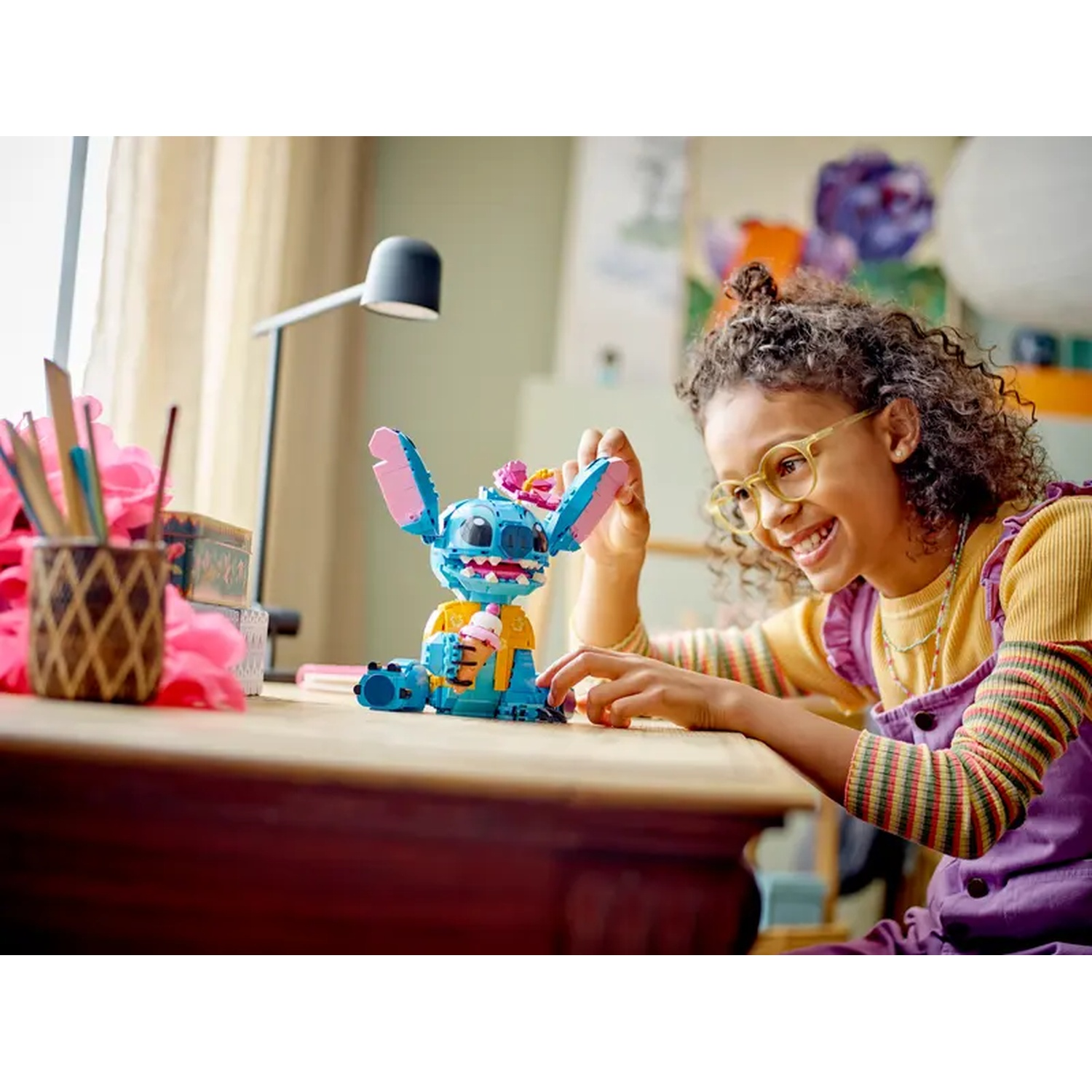 Een meisje glimlacht naar een Stitch Lego-figuur. De figuur houdt ijs vast. Het meisje draagt een bril en een paarse overall.