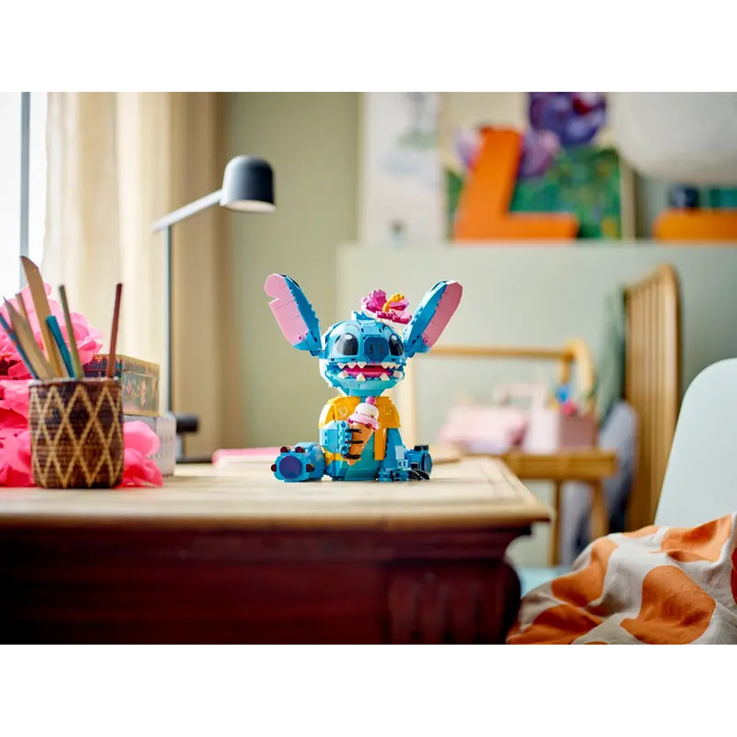 Een Lego Stitch figuur met ijs zit op een bureau. Een lamp, bloemen en kunstwerken staan op de achtergrond. Rechts staat een stoel.
