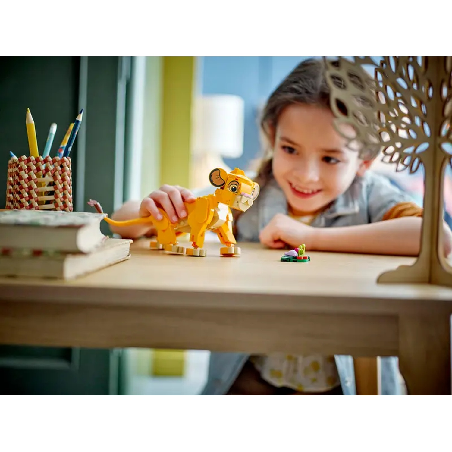 Une enfant sourit à un lion Lego sur une table. Des crayons, des livres et une décoration d'arbre en bois sont également présents.