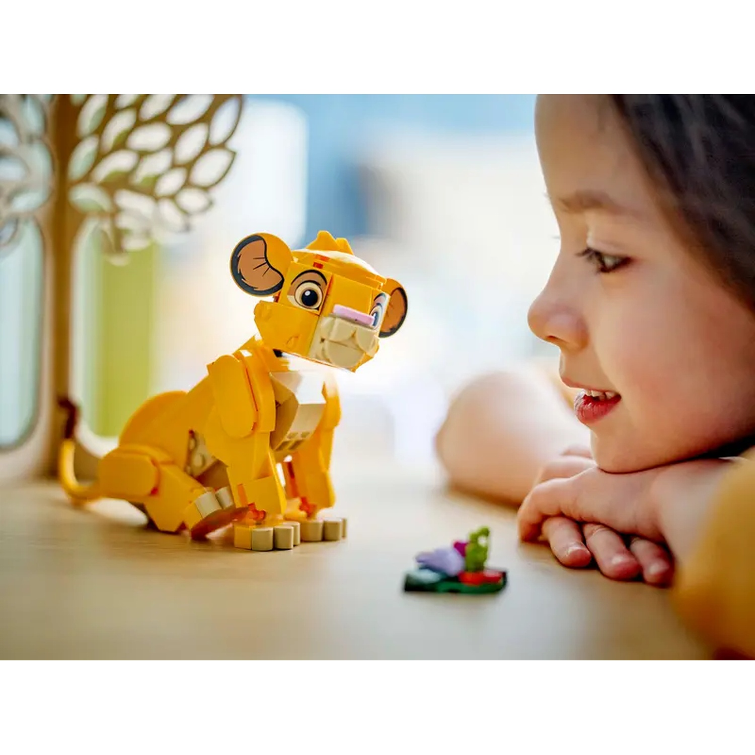 Une fille regarde un jouet Lego Simba. Le lion jaune est assis sur une table près d'un petit objet Lego. Une forme d'arbre est en arrière-plan.