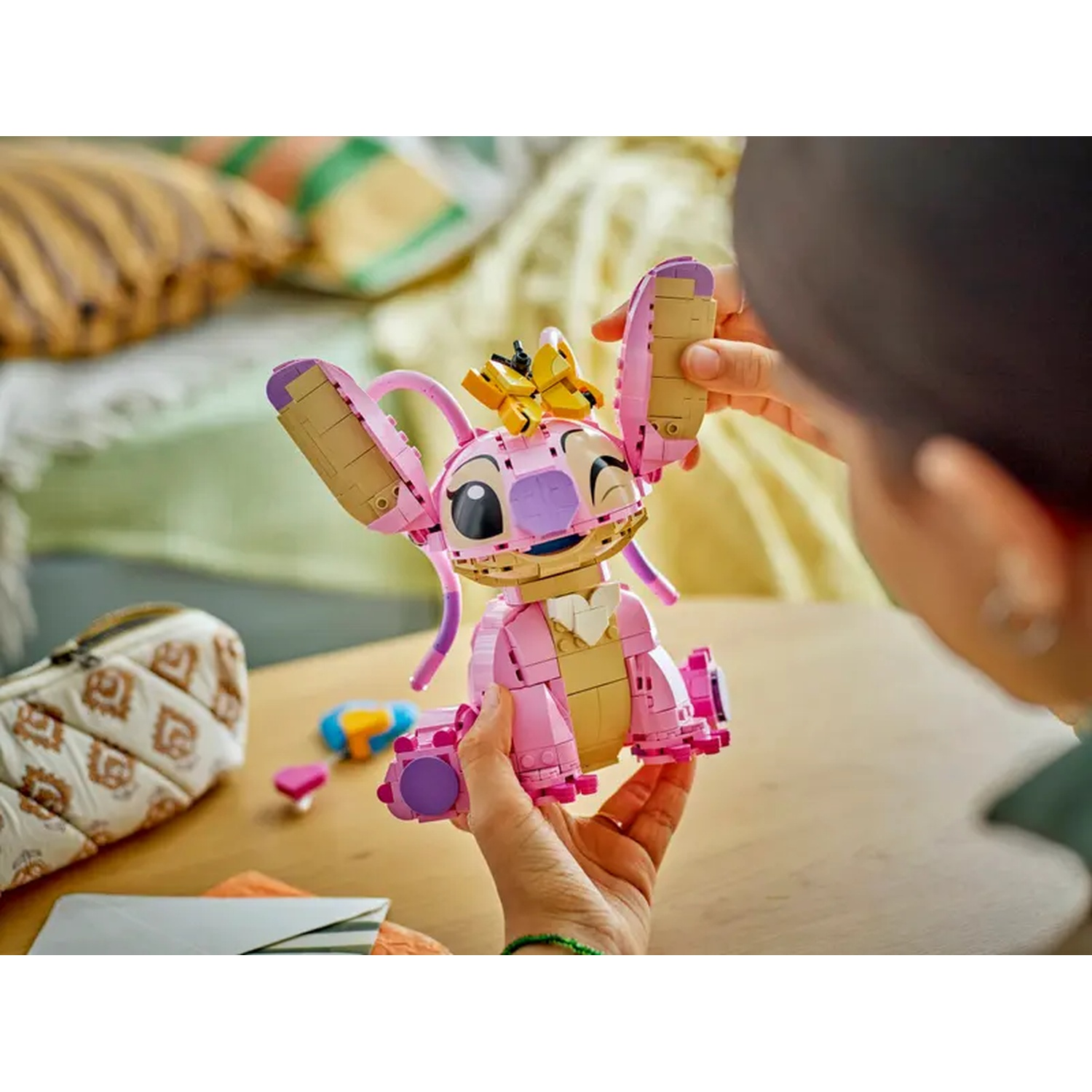 Een persoon houdt een roze Lego Stitch figuur met een kroon vast. Andere Lego stukken, een etui en kussens op de achtergrond.