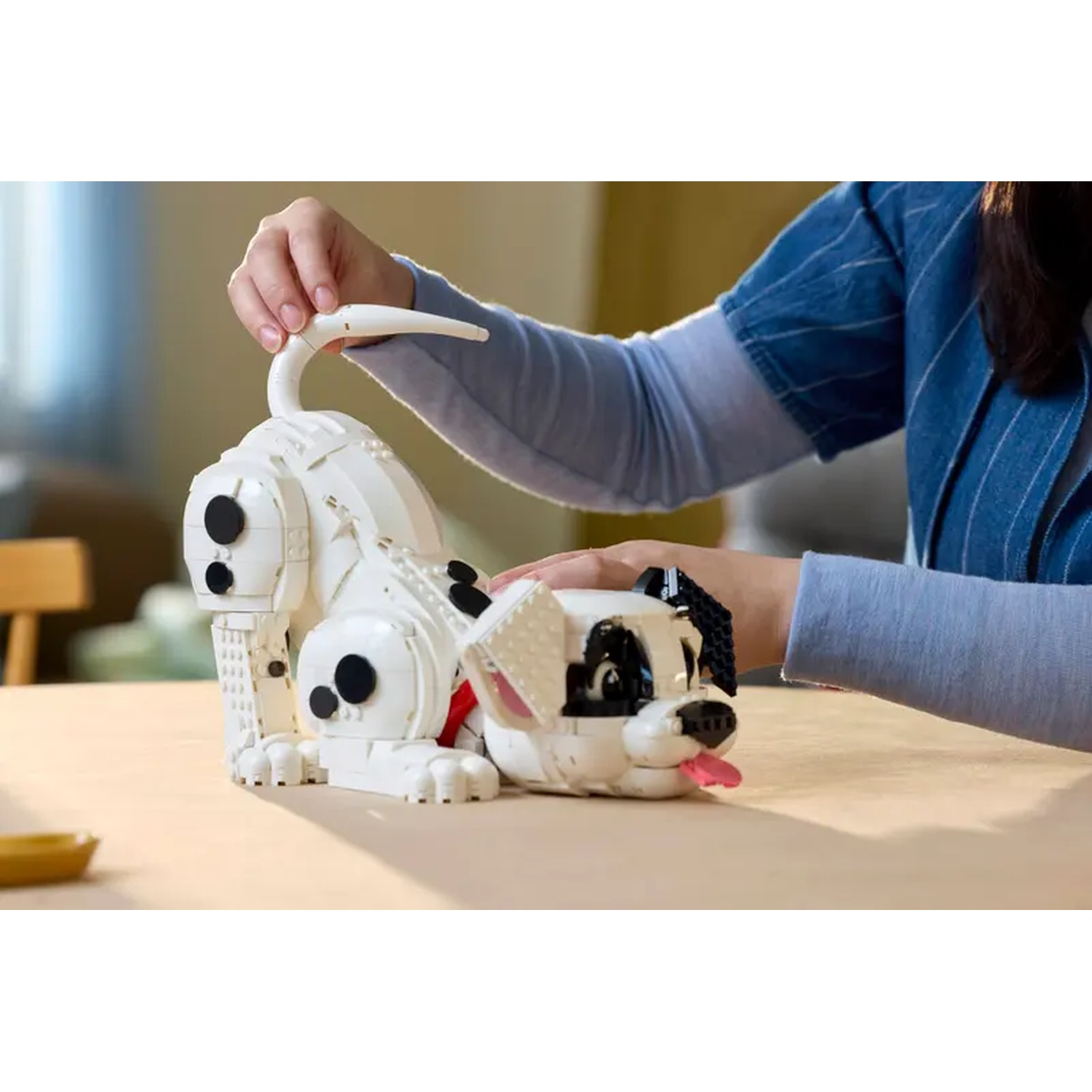 Een persoon assembleert een witte Lego Dalmatische hond met zwarte vlekken. De hond heeft een roze tong. Een tafel en armen zijn zichtbaar.