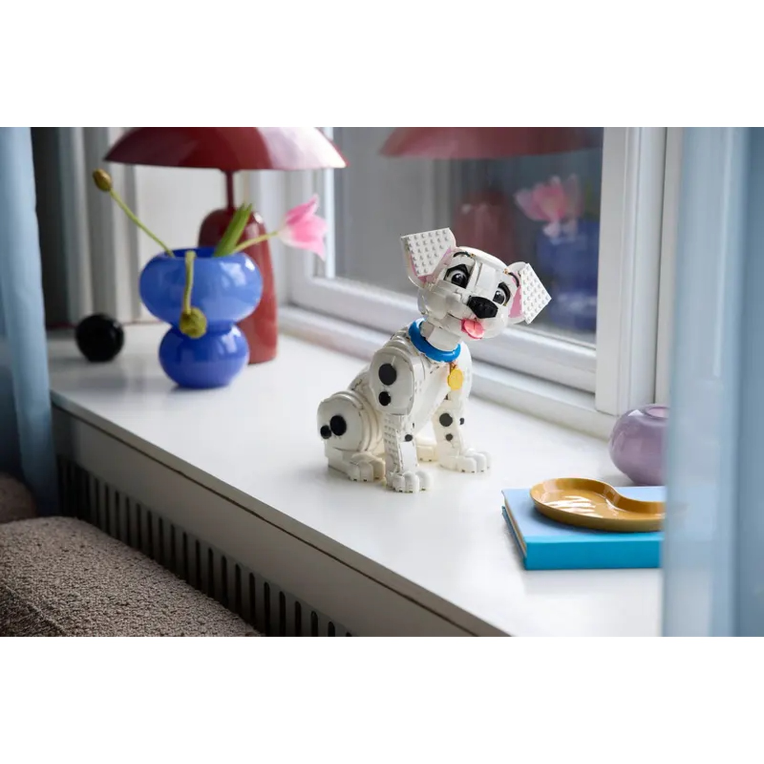 Een Lego Dalmatische hond zit op een witte vensterbank. Bloemen in een blauwe vaas staan op de achtergrond, samen met een rode lamp en een blauw boek.