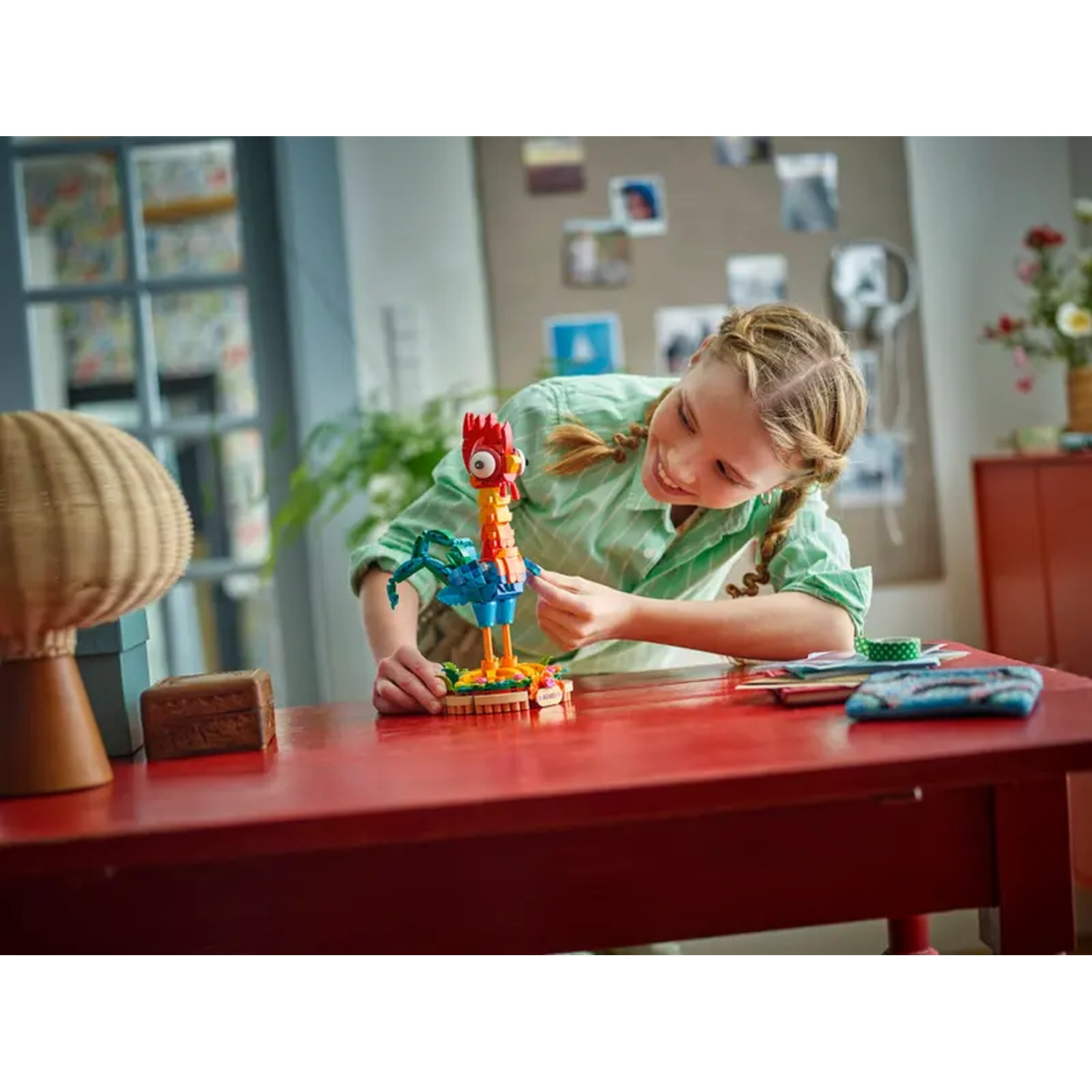 A girl builds a Lego chicken on a red table. There is a woven lamp and a wooden box. The background shows a room.