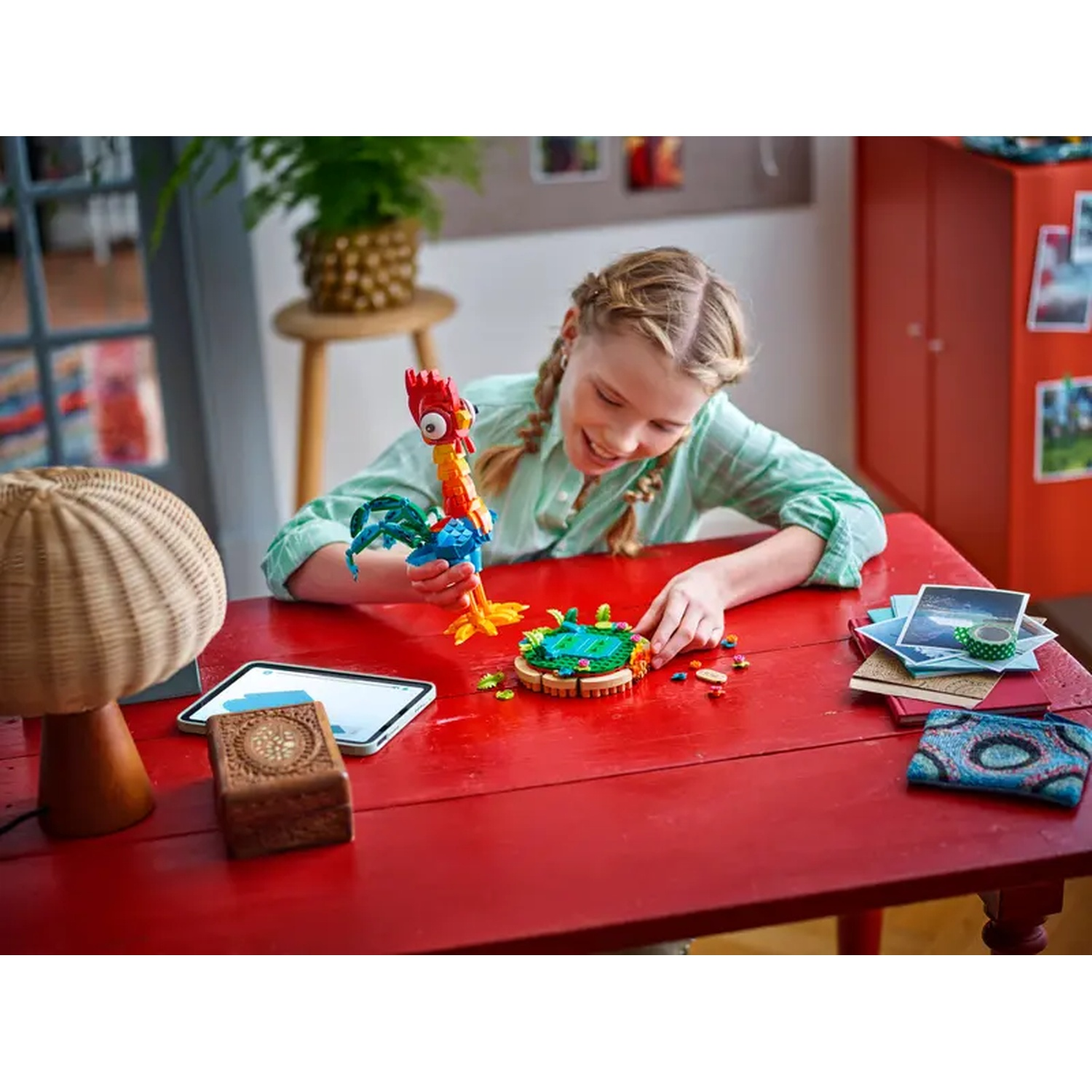 A girl smiles while building a Lego set on a red table. A rooster Lego figure is in her hand.
