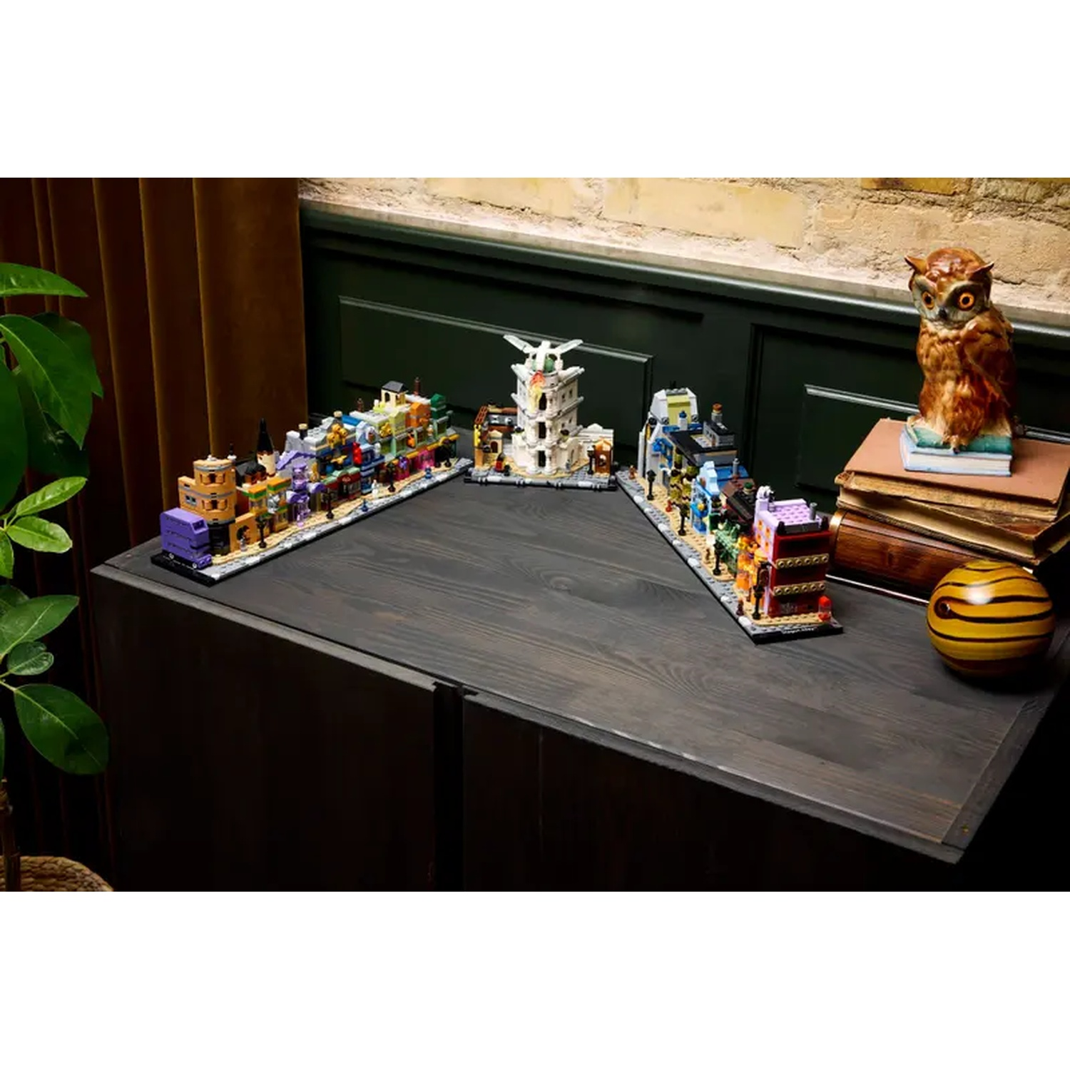 A dark wooden table displays Lego buildings. An owl statue sits on books. A striped sphere is on the table. Green plant on the left.