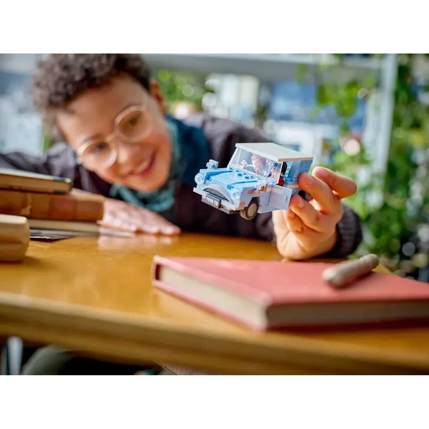 Une personne tient une voiture jouet bleue. Un livre rouge et d'autres objets sont sur une table en bois. La personne porte des lunettes.
