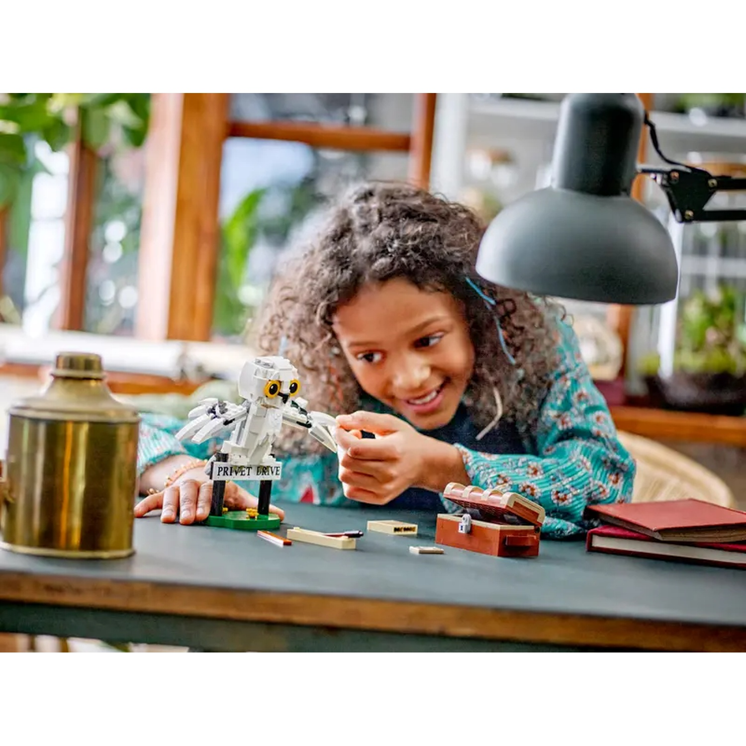 A girl smiles while assembling a Lego owl. A lamp, chest, books, and a golden container are on the table.