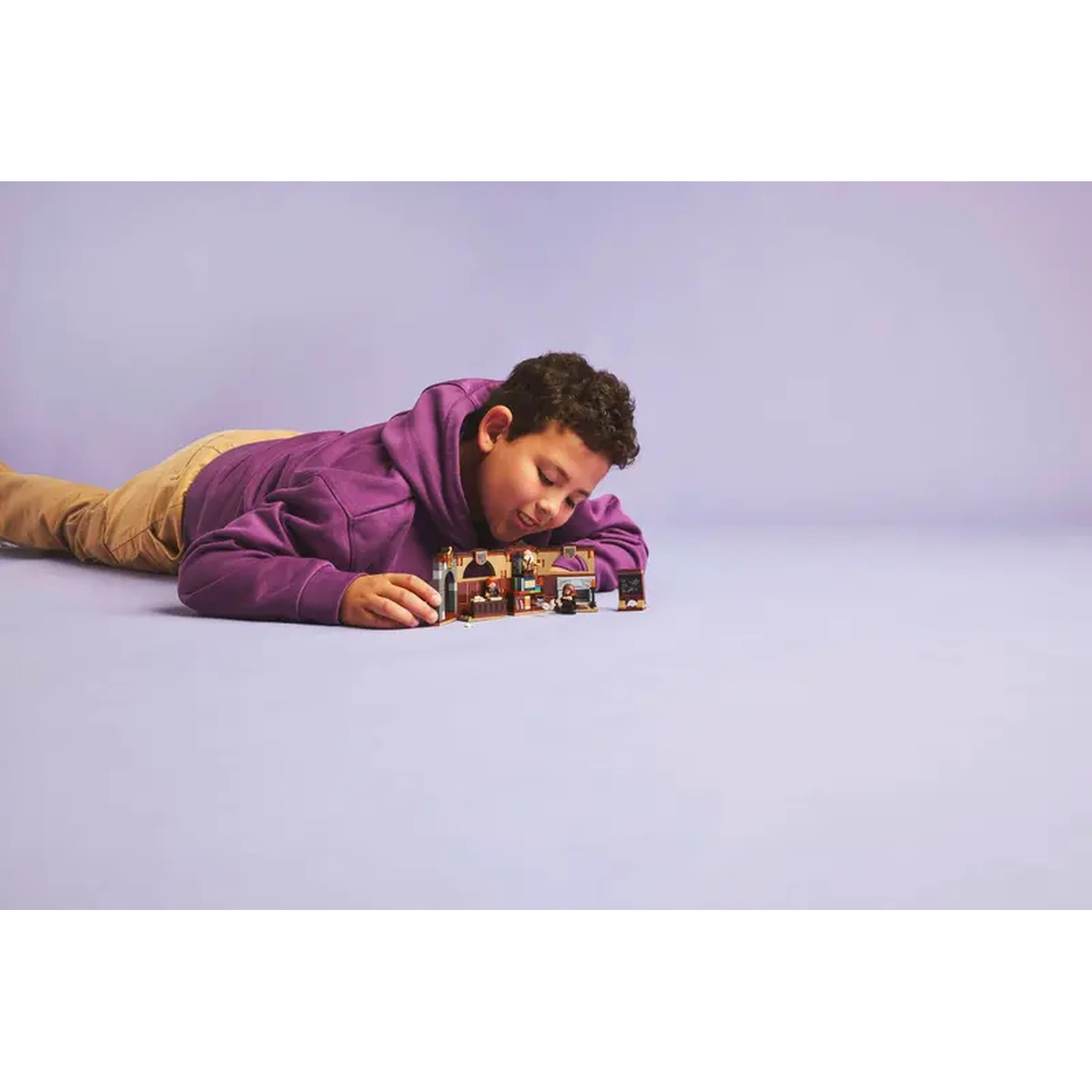 A boy in a purple hoodie and khaki pants lies on a purple surface, looking at a Lego set. The set has multiple small buildings.