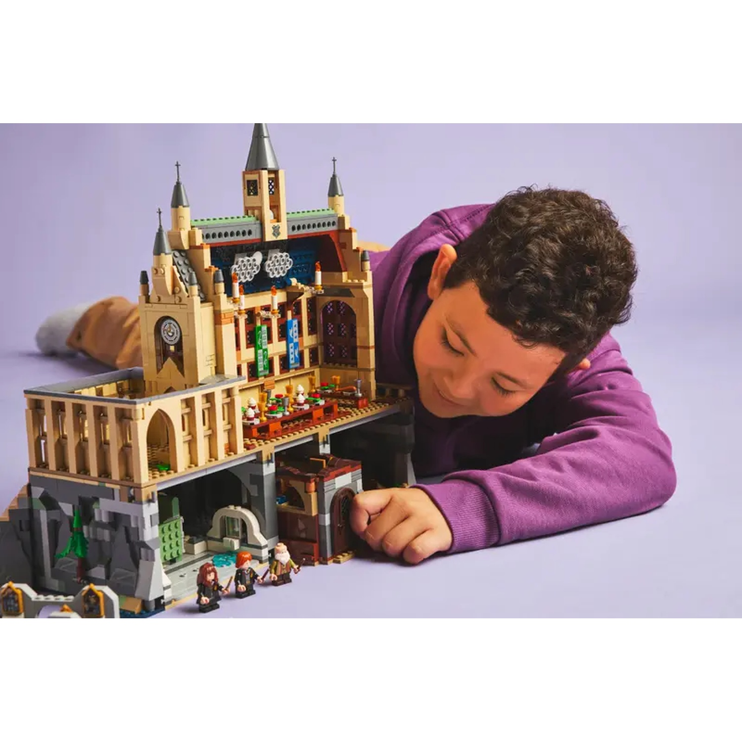 A boy lies on a purple surface, looking at a Lego castle. The castle is tan and gray with small figures.