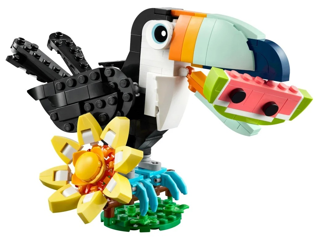 Lego tukan trzyma kawałek arbuza. Siedzi na podstawie z kwiatów i trawy. Ptak jest czarny, biały i niebieski.