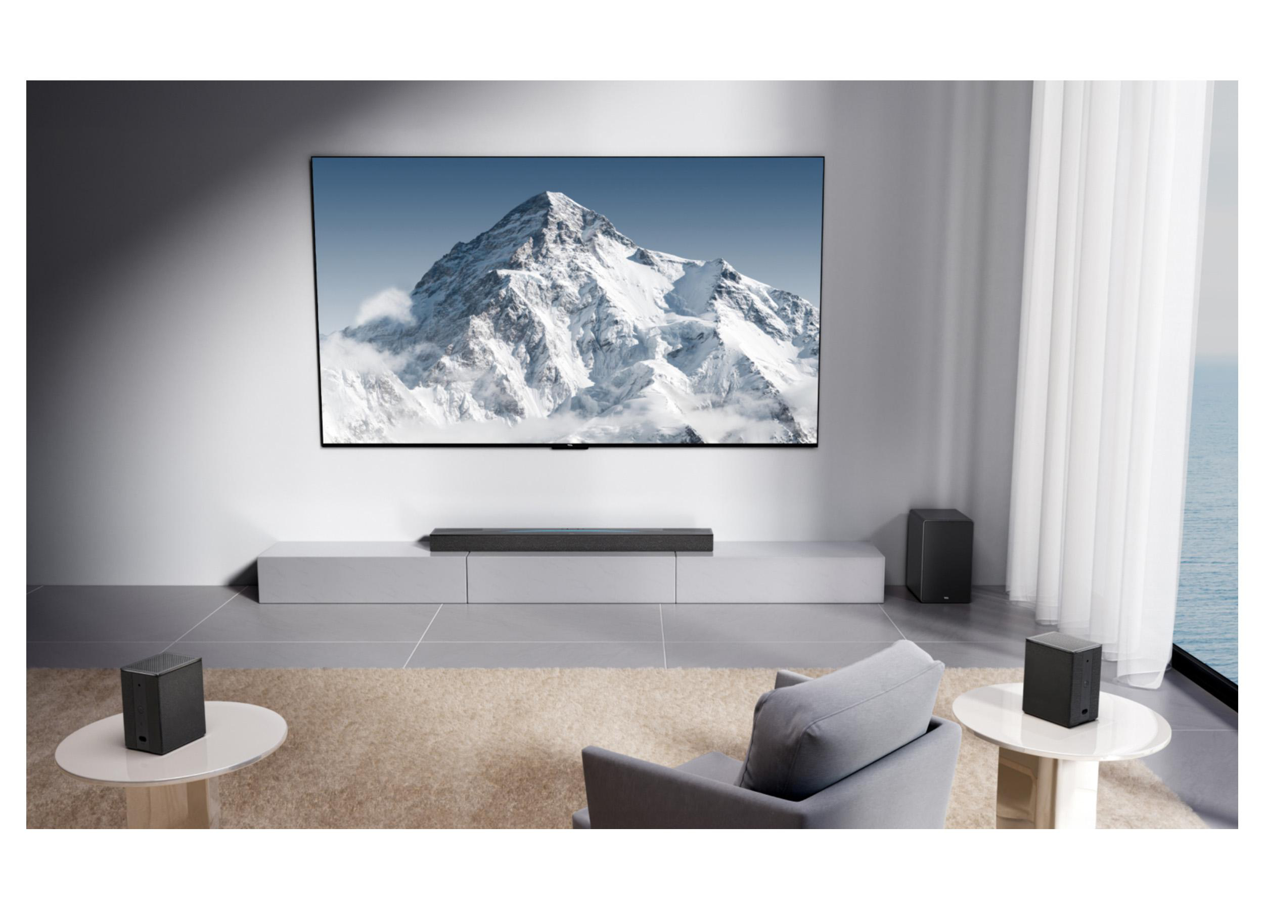 Wohnzimmer mit TV, der einen Berg zeigt. Soundbar, Lautsprecher, grauer Stuhl und Tische. Meerblick durch das Fenster.