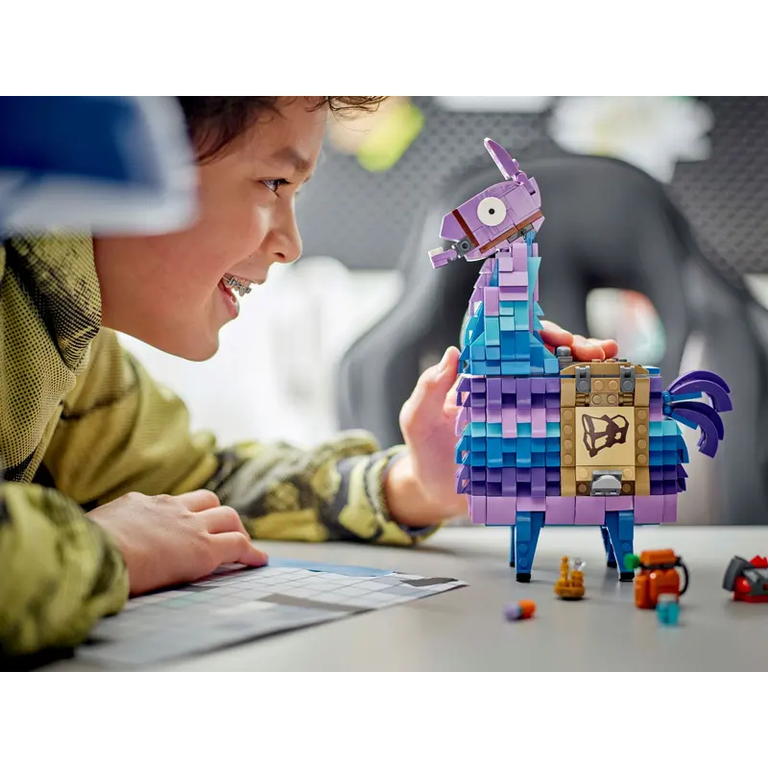 Een jongen met een beugel lacht, kijkend naar een Lego-lama. De lama is paars, blauw en bruin. Kleine Lego-stukjes liggen op tafel.
