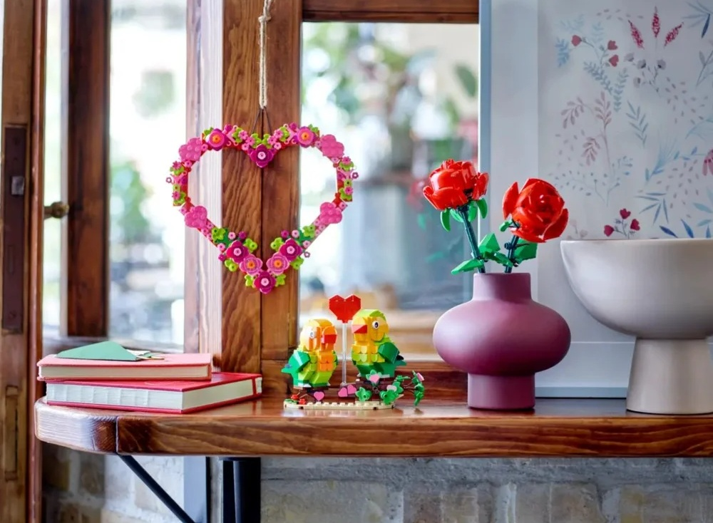 Drewniana półka z dekoracjami Lego: wianek w kształcie serca, róże w wazonie, papugi i książki. Światło z okna.