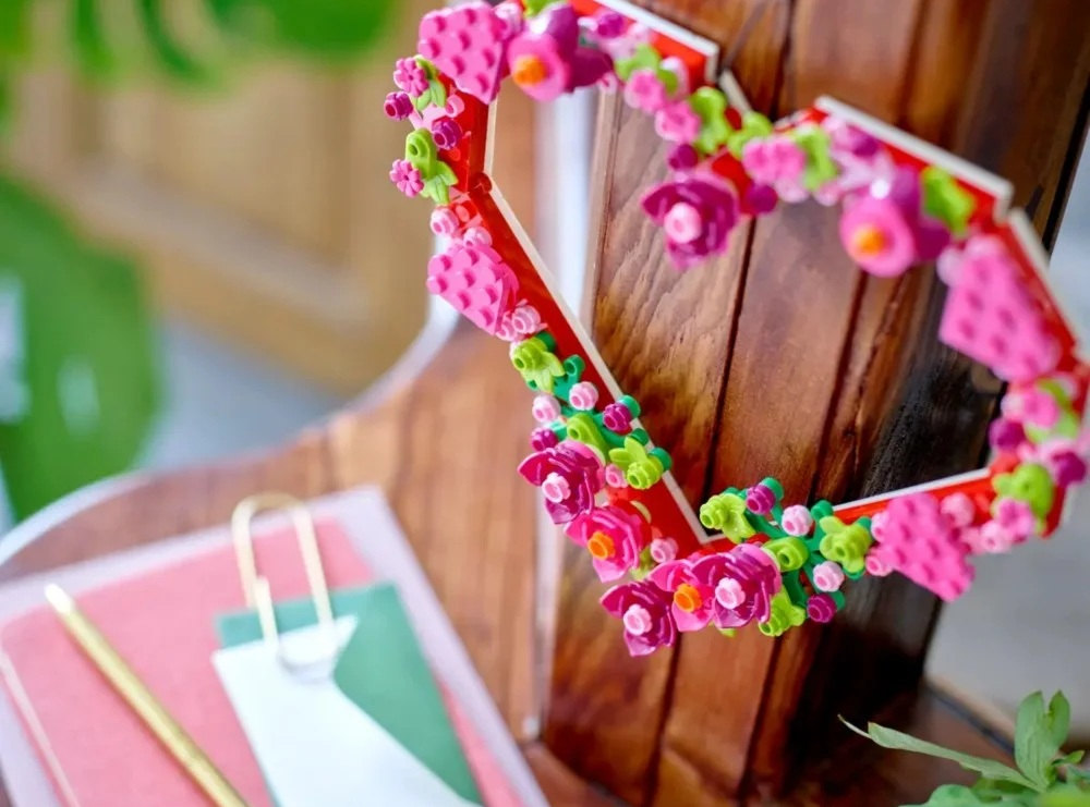 Ozdoba w kształcie serca z różowych kwiatów i czerwonej ramy, na drewnianej powierzchni, z notatnikiem i długopisem w tle.