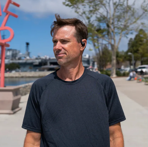 Mężczyzna w ciemnej koszulce i słuchawkach stoi na zewnątrz w pobliżu nabrzeża. Tło obejmuje statek, drzewa i błękitne niebo.