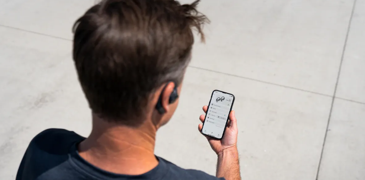 Mężczyzna patrzy na swój smartfon, nosząc słuchawki. Jest na zewnątrz, na betonowej powierzchni. Telefon wyświetla interfejs aplikacji.