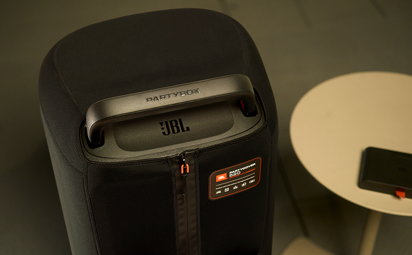 Schwarzer JBL PartyBox-Lautsprecher auf einem Tisch. Griff, Logo und Reißverschluss sind sichtbar. Ein kleiner weißer Tisch und ein schwarzes Gerät sind in der Nähe.