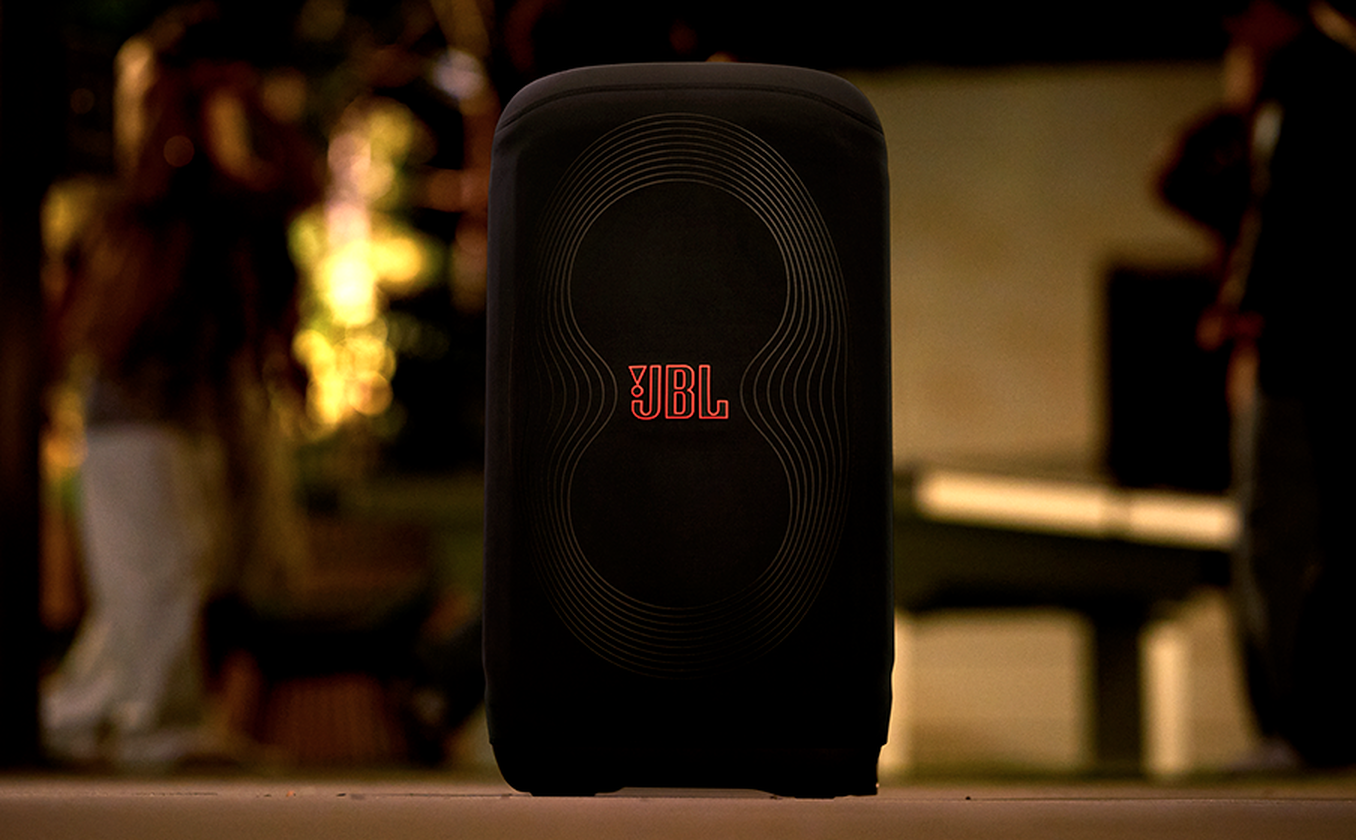 Schwarzer JBL-Lautsprecher mit rotem Logo, Personen im Hintergrund verschwommen. Lautsprecher hat wellenartiges Design.