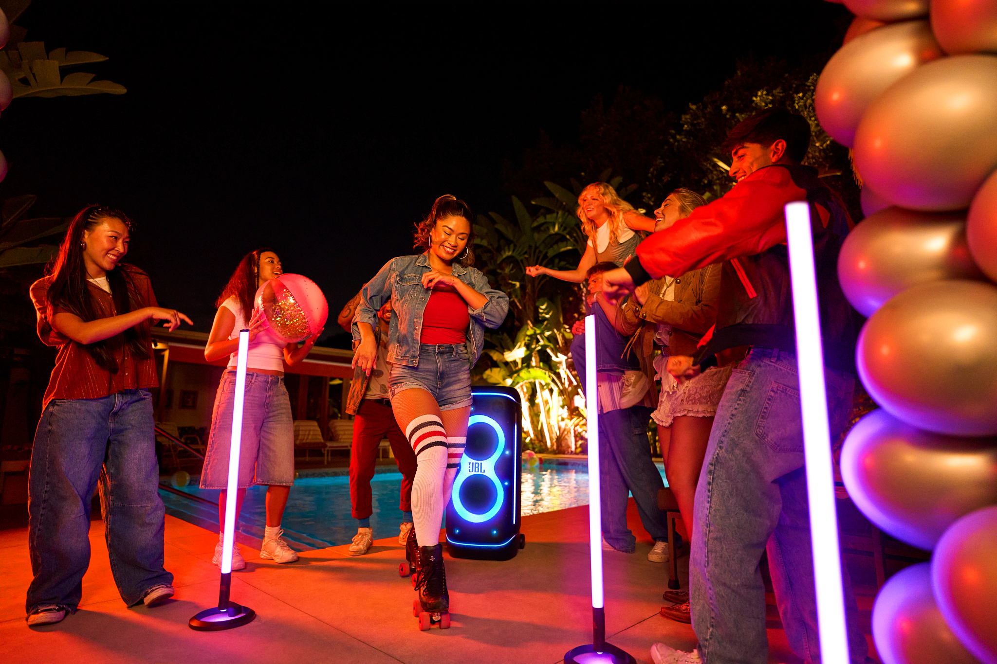 Des gens dansent à une fête nocturne. Une femme en patins à roulettes est au centre. Une enceinte JBL est à proximité.