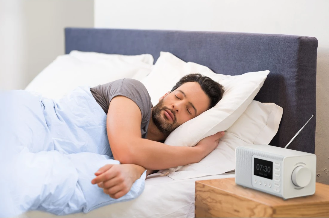 Un homme dort dans son lit. Un radio-réveil blanc est sur une table de chevet en bois. L'heure affichée est 07h30.