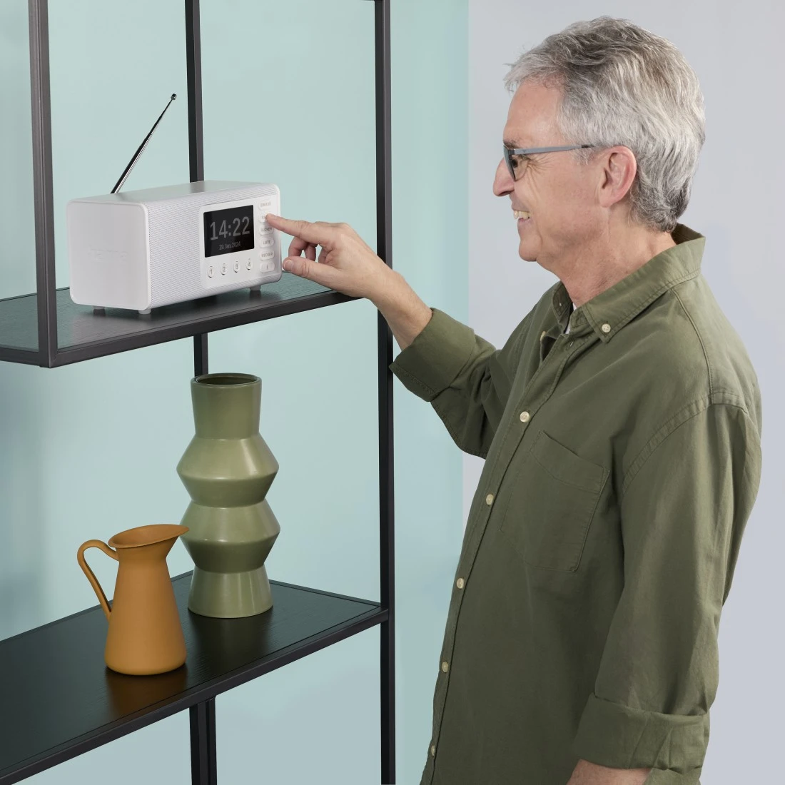 Un homme âgé portant des lunettes règle une radio blanche sur une étagère. Il y a des vases en dessous. Le fond est bleu clair et blanc.