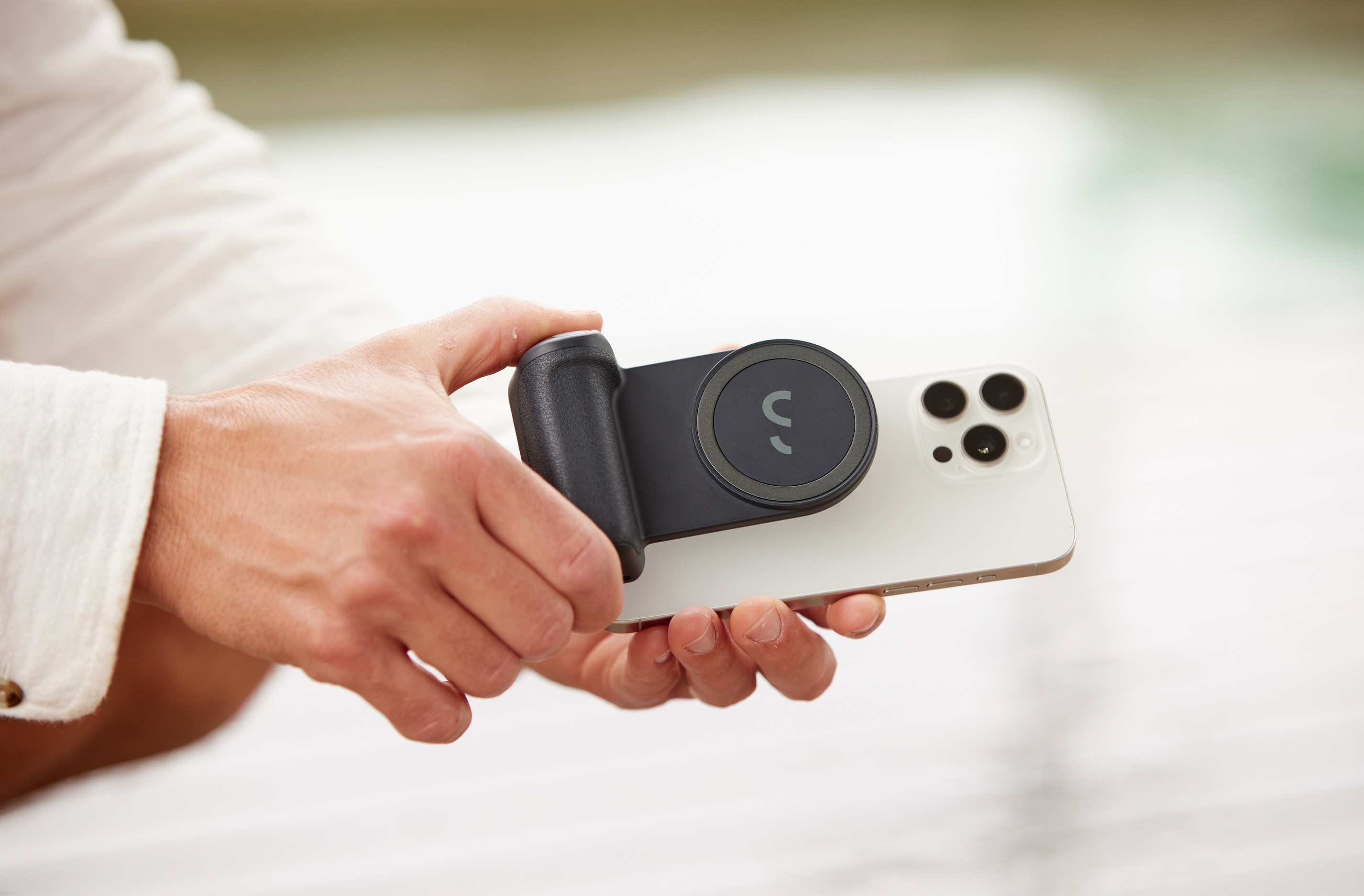 Mani che tengono un telefono con un accessorio nero attaccato. Lo sfondo ha un effetto sfocato verde e bianco.