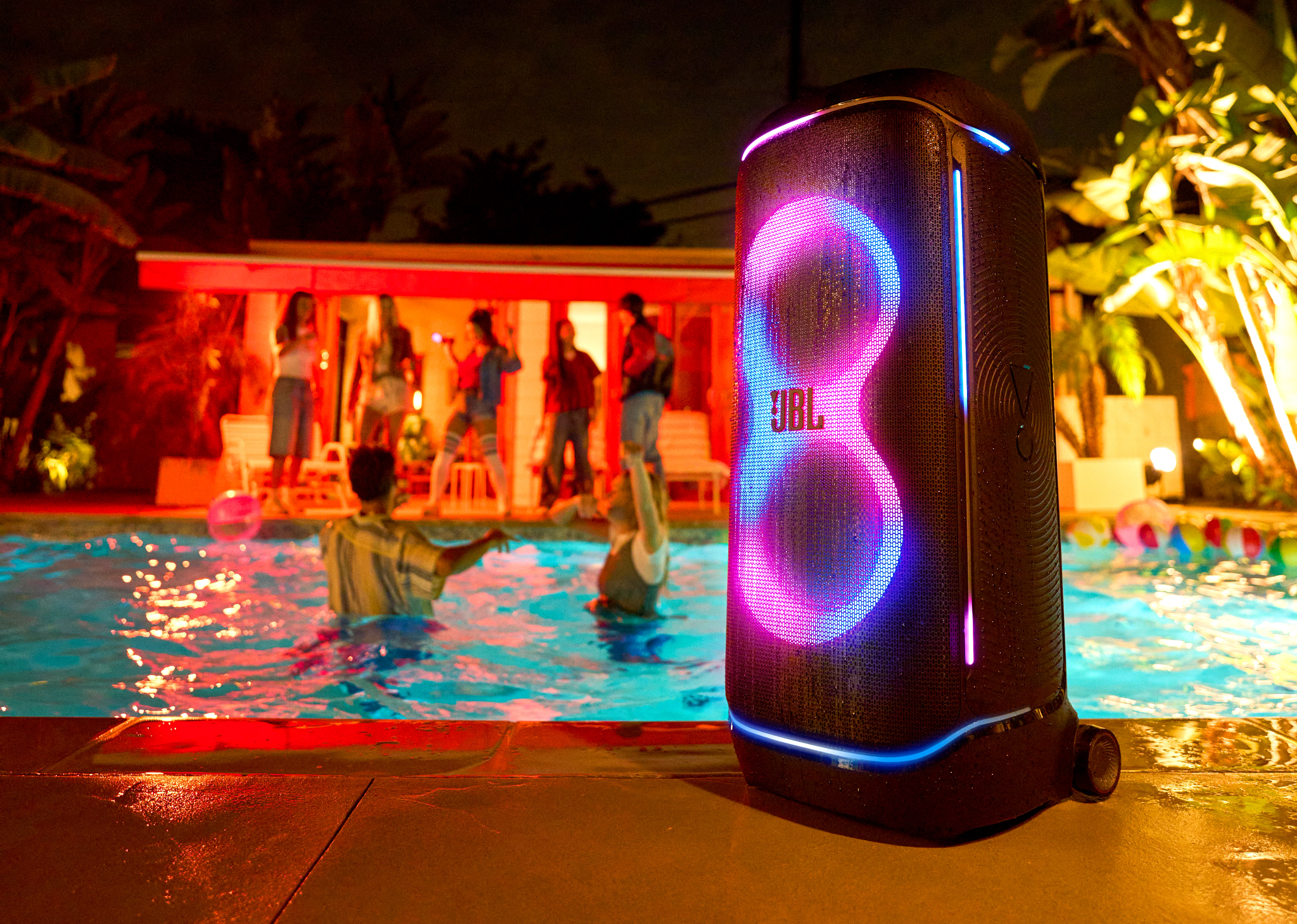 Scène de fête nocturne avec piscine. Un haut-parleur JBL brille. Des gens dansent près d'une piscine. Palmiers et bâtiment en arrière-plan.
