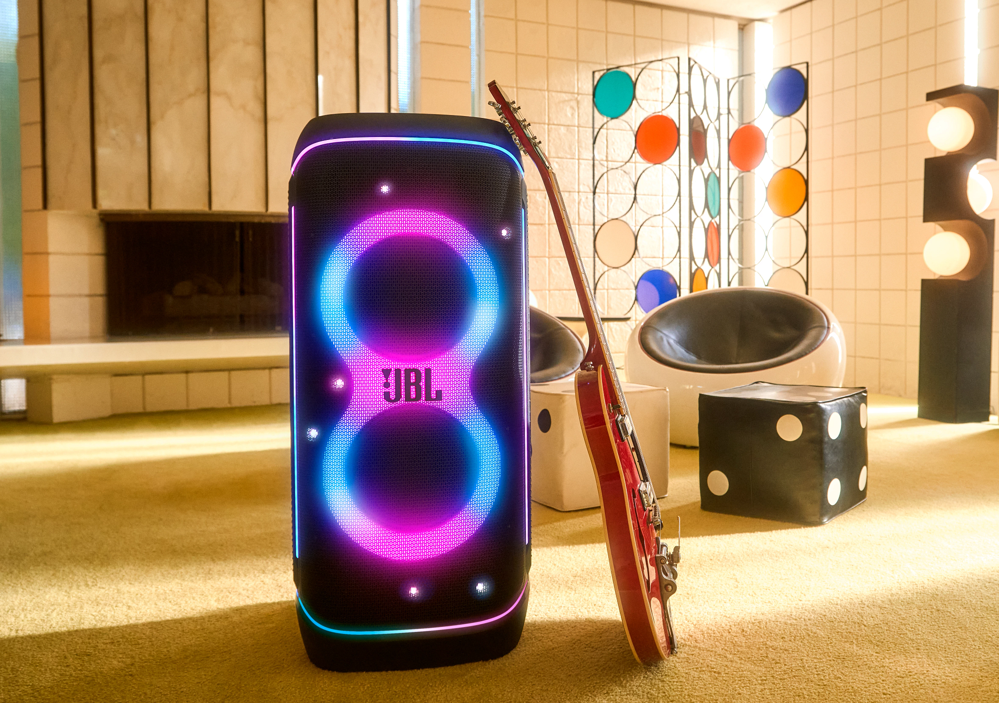 Un haut-parleur JBL brille en bleu et rose. Une guitare rouge s'appuie dessus. Un pouf en forme de dé est à proximité.