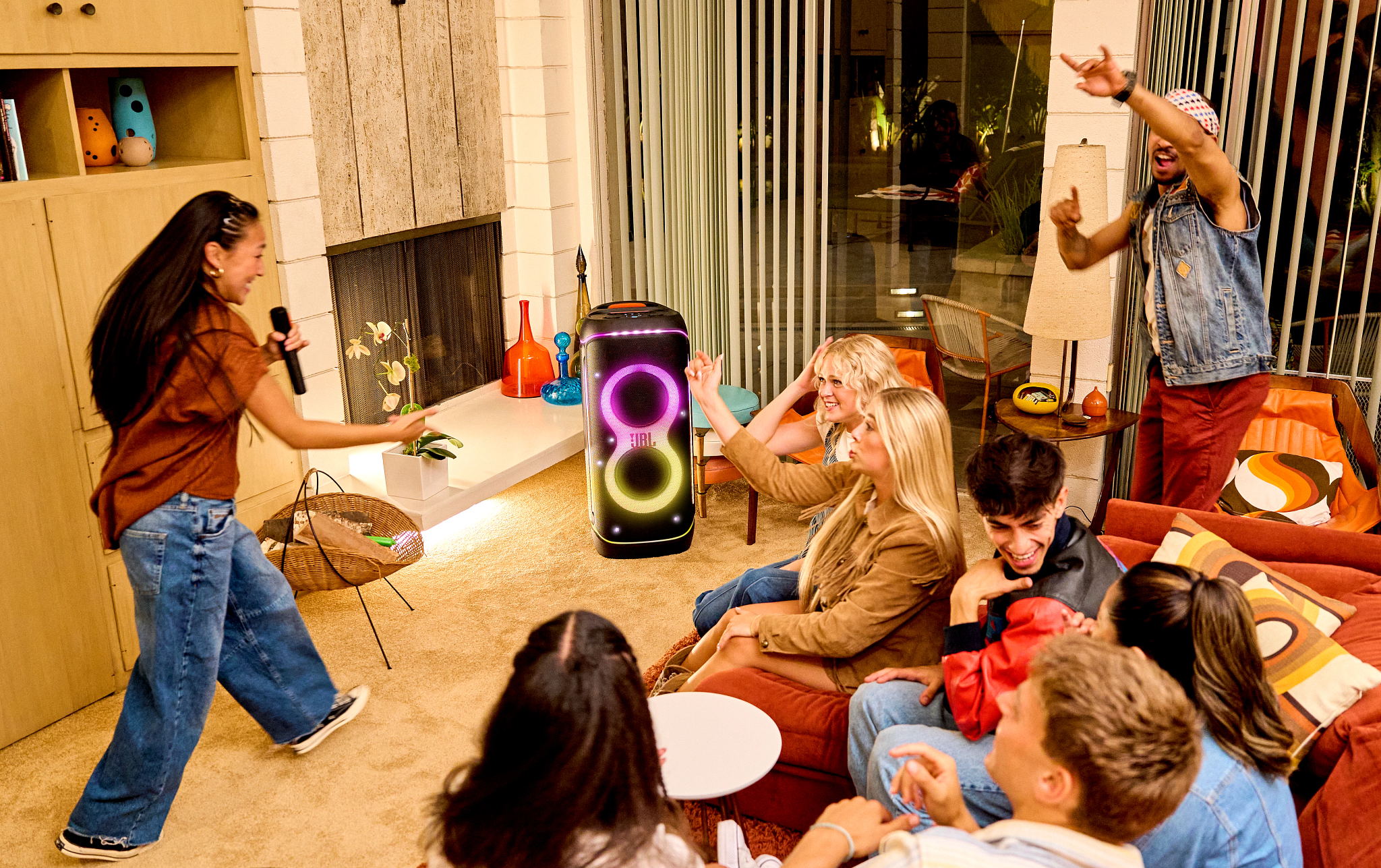 Un groupe de personnes est à une fête, une femme tient un microphone, un homme danse et une enceinte est en arrière-plan.