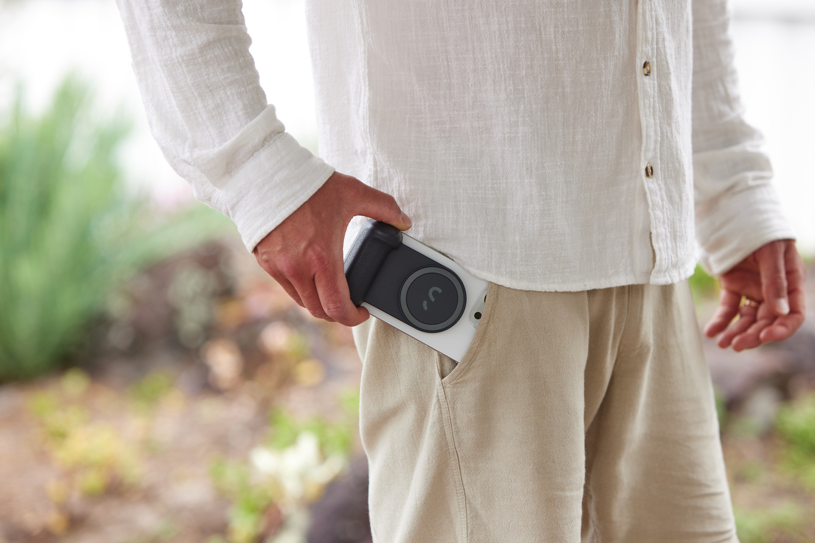 Una persona con camicia bianca e pantaloncini beige tiene in mano un telefono con un accessorio nero in tasca.