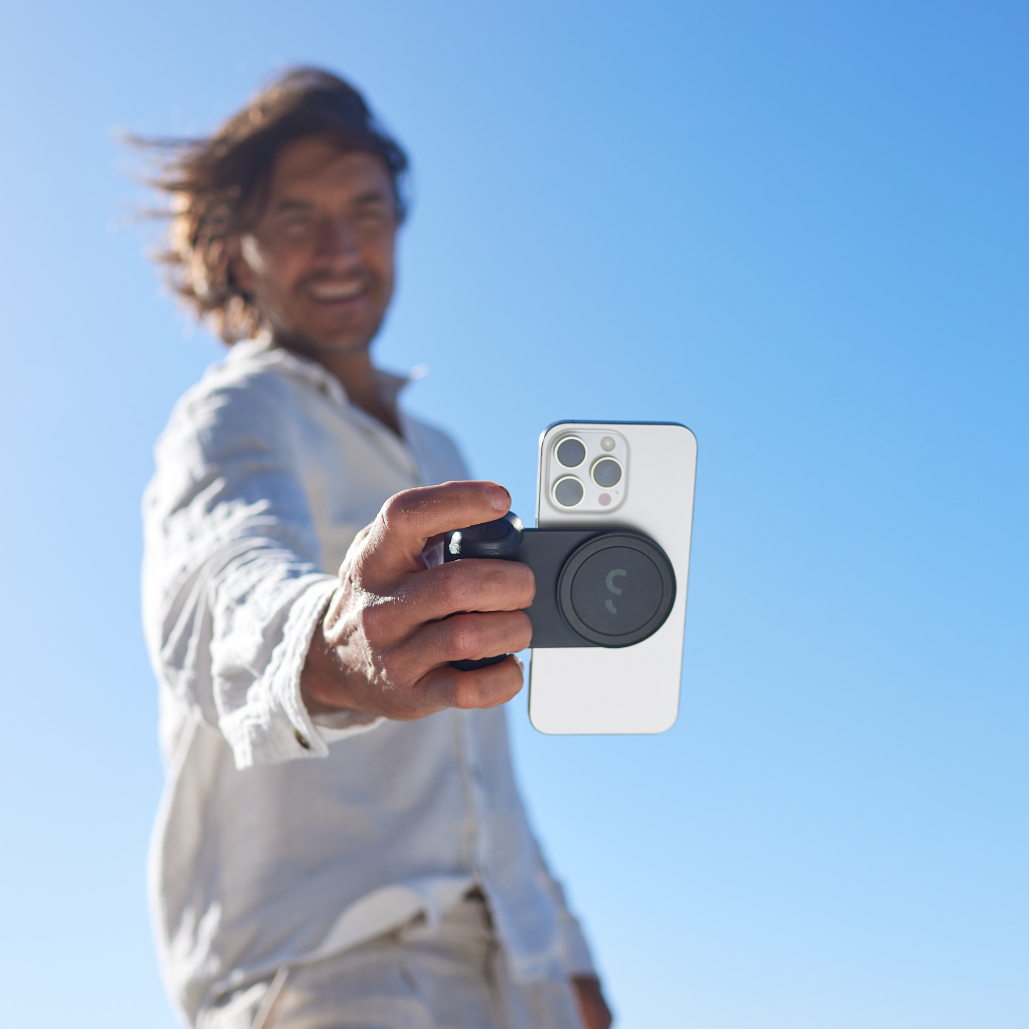 Uomo che tiene un telefono con impugnatura magnetica, contro un cielo azzurro. Sta sorridendo. Il telefono è bianco.