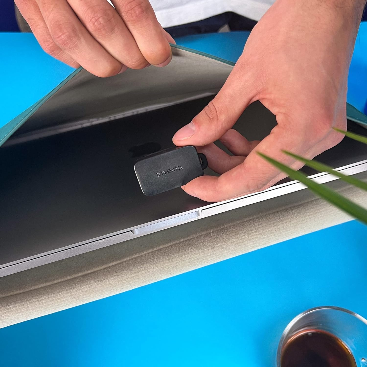 Mani che inseriscono un dispositivo nero in una custodia per laptop. Il laptop è su una superficie blu, con un bicchiere di caffè nell'angolo.