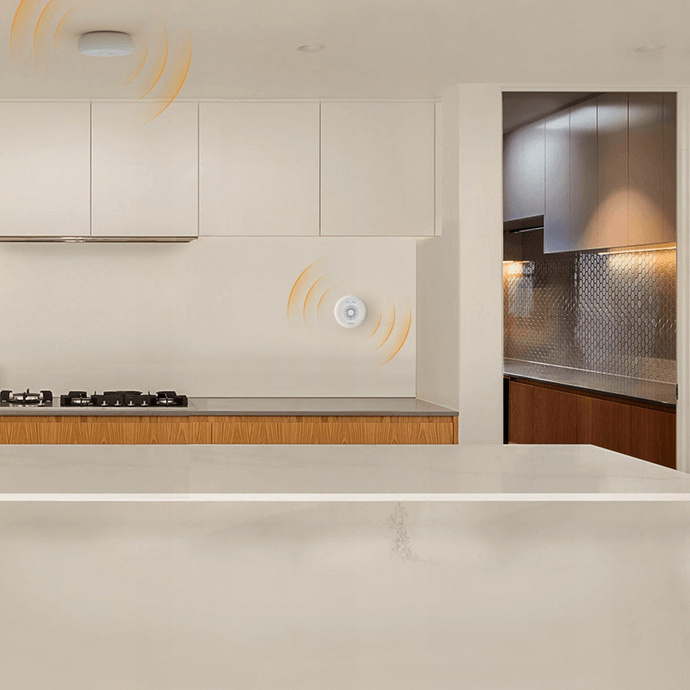 Wnętrze kuchni z czujnikiem dymu na ścianie. Widoczne szafki i kuchenka. Otwarte drzwi prowadzą do innego pokoju.